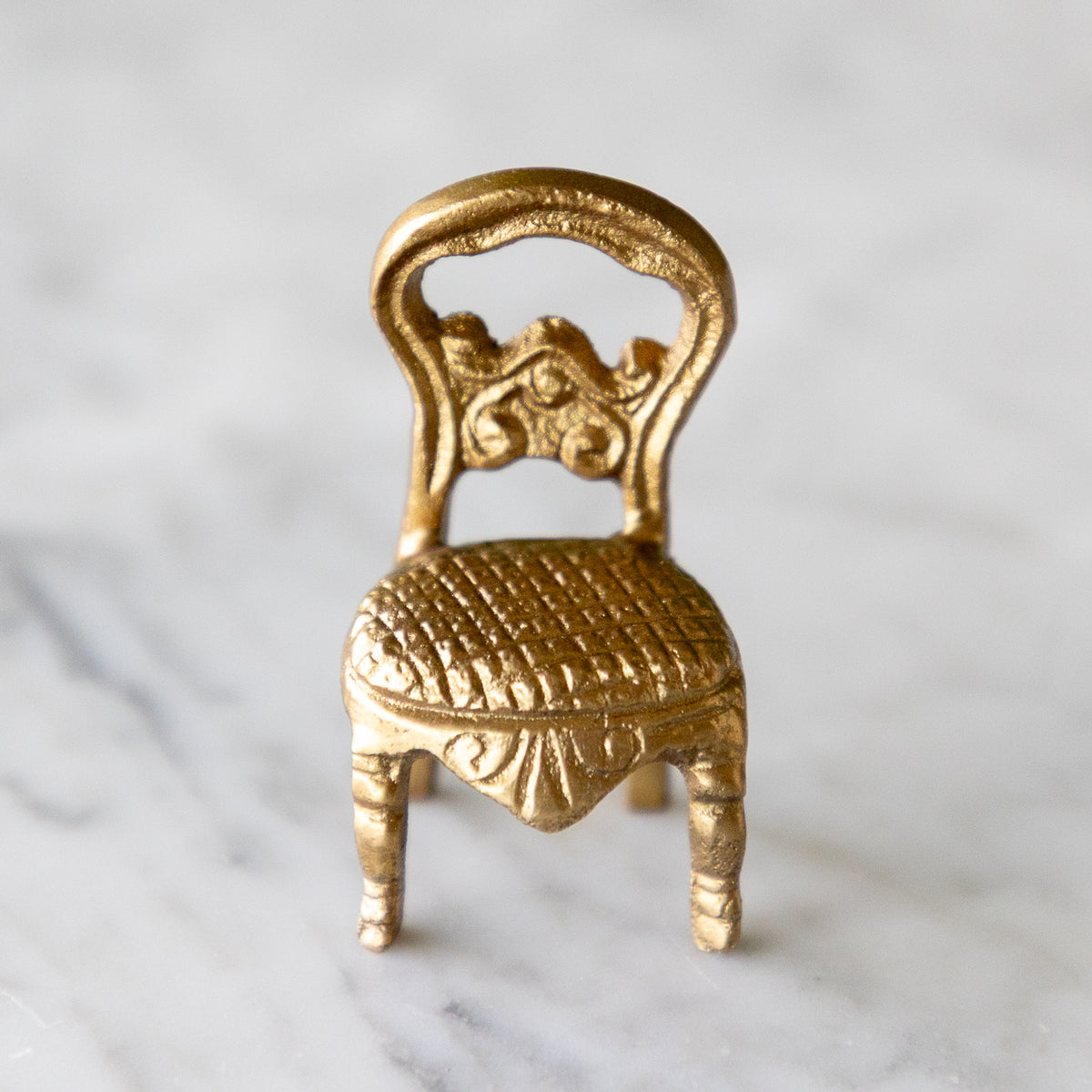 A miniature gold-finished chair figurine with an ornate backrest and textured seat, displayed on a light marble surface.