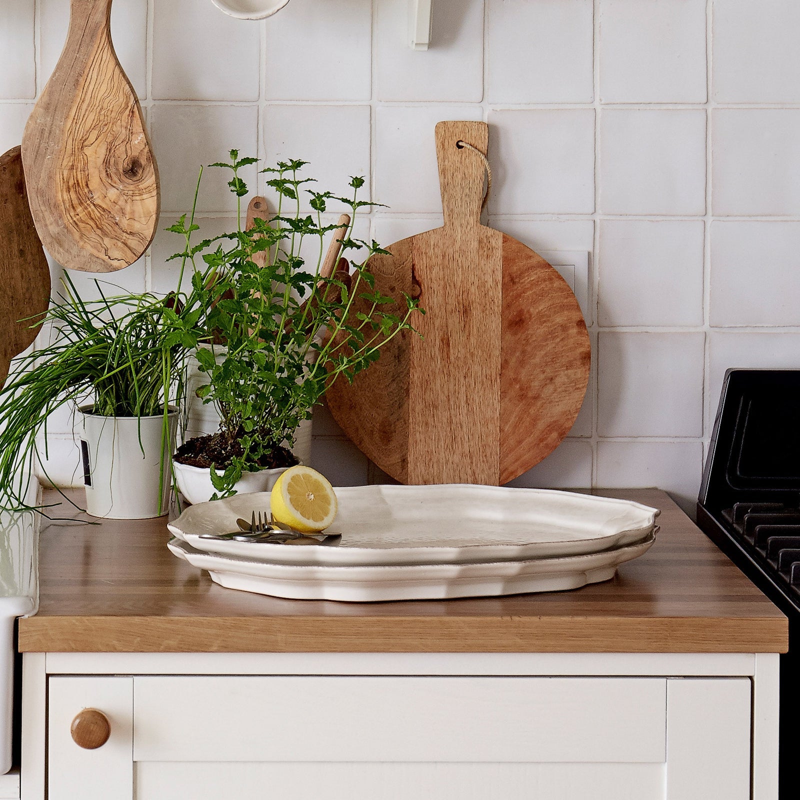 Kitchen counter with wooden cutting boards, a white platter, and a lemon on a tiled wall background.