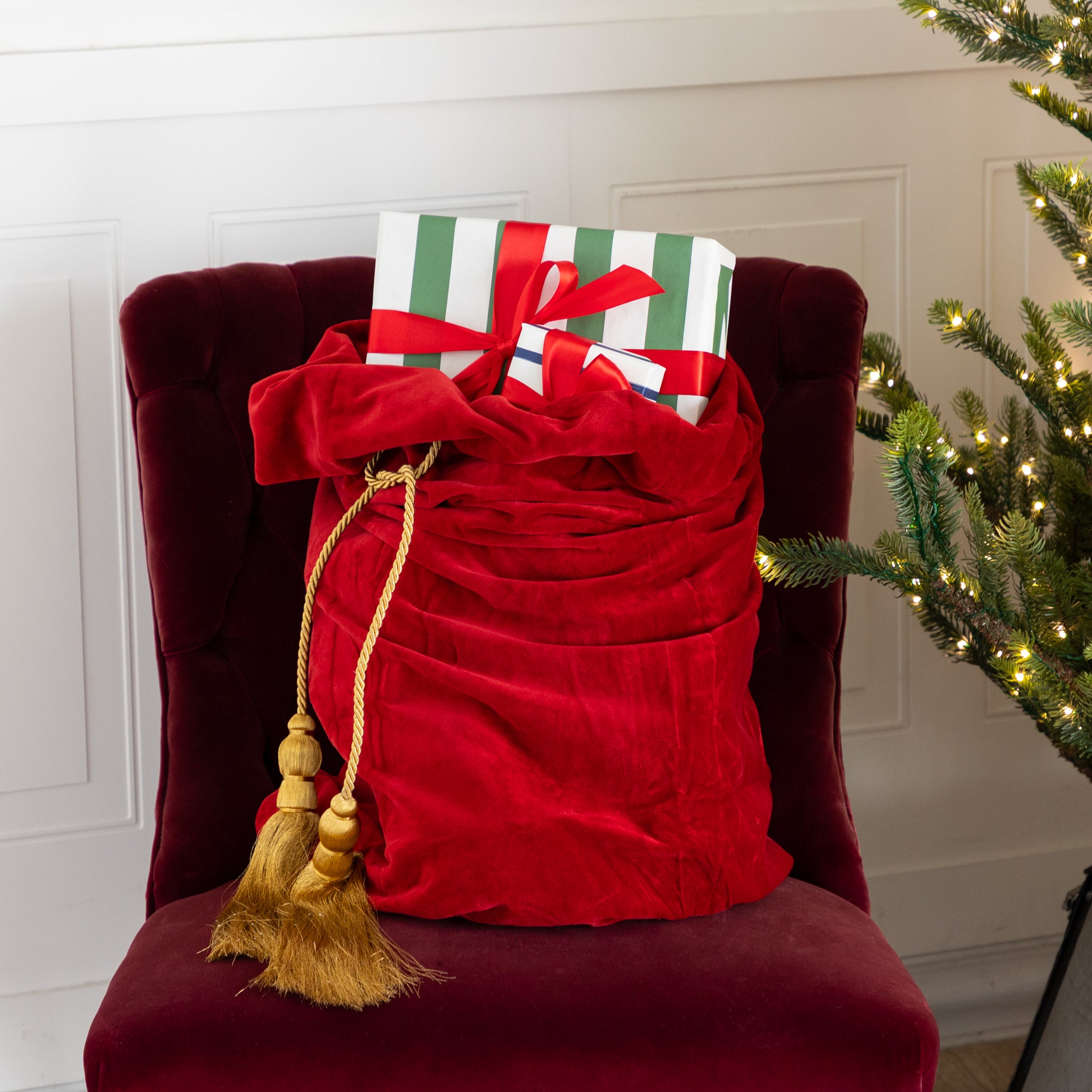 Red Velvet Santa Bag with two gold tassels and gifts inside, on a red chair next to a decorated Christmas tree.