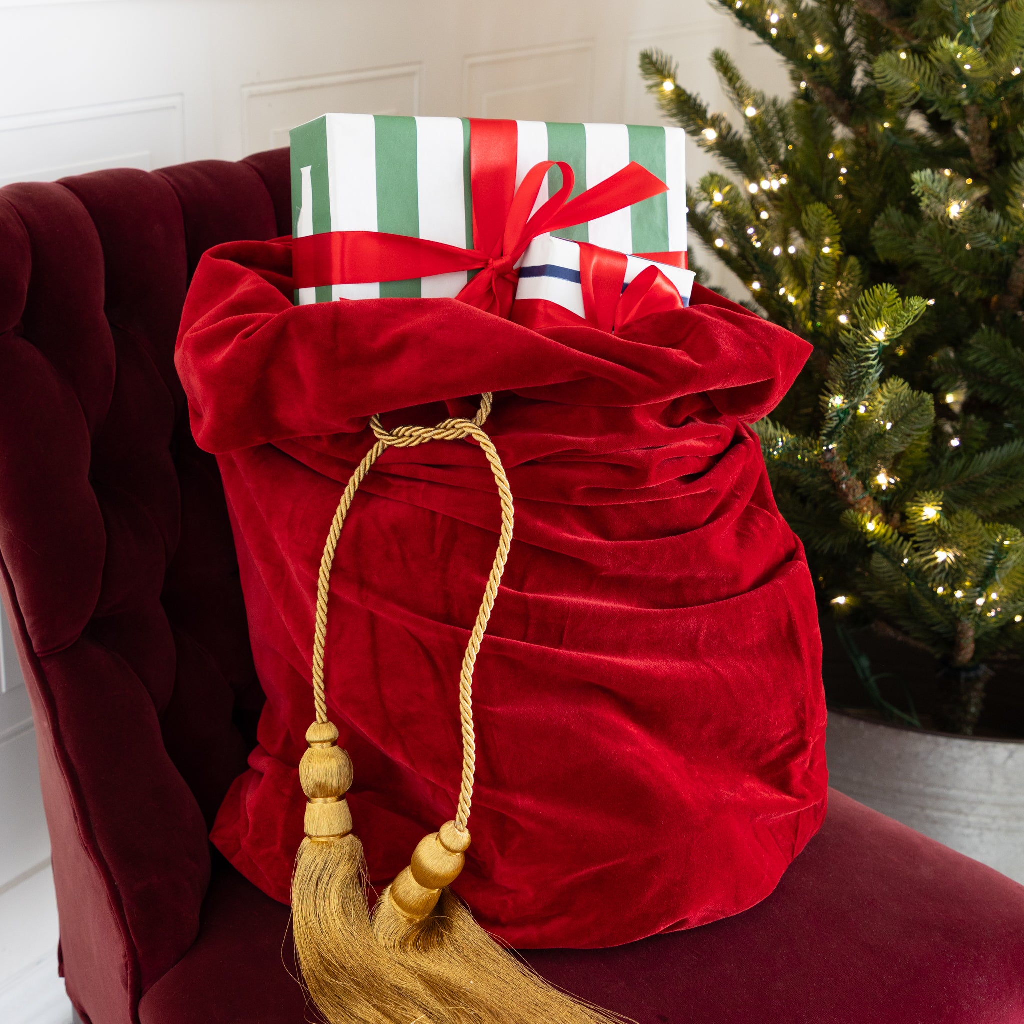 Red Velvet Santa Bag with two gold tassels and gifts inside, on a red chair next to a decorated Christmas tree.