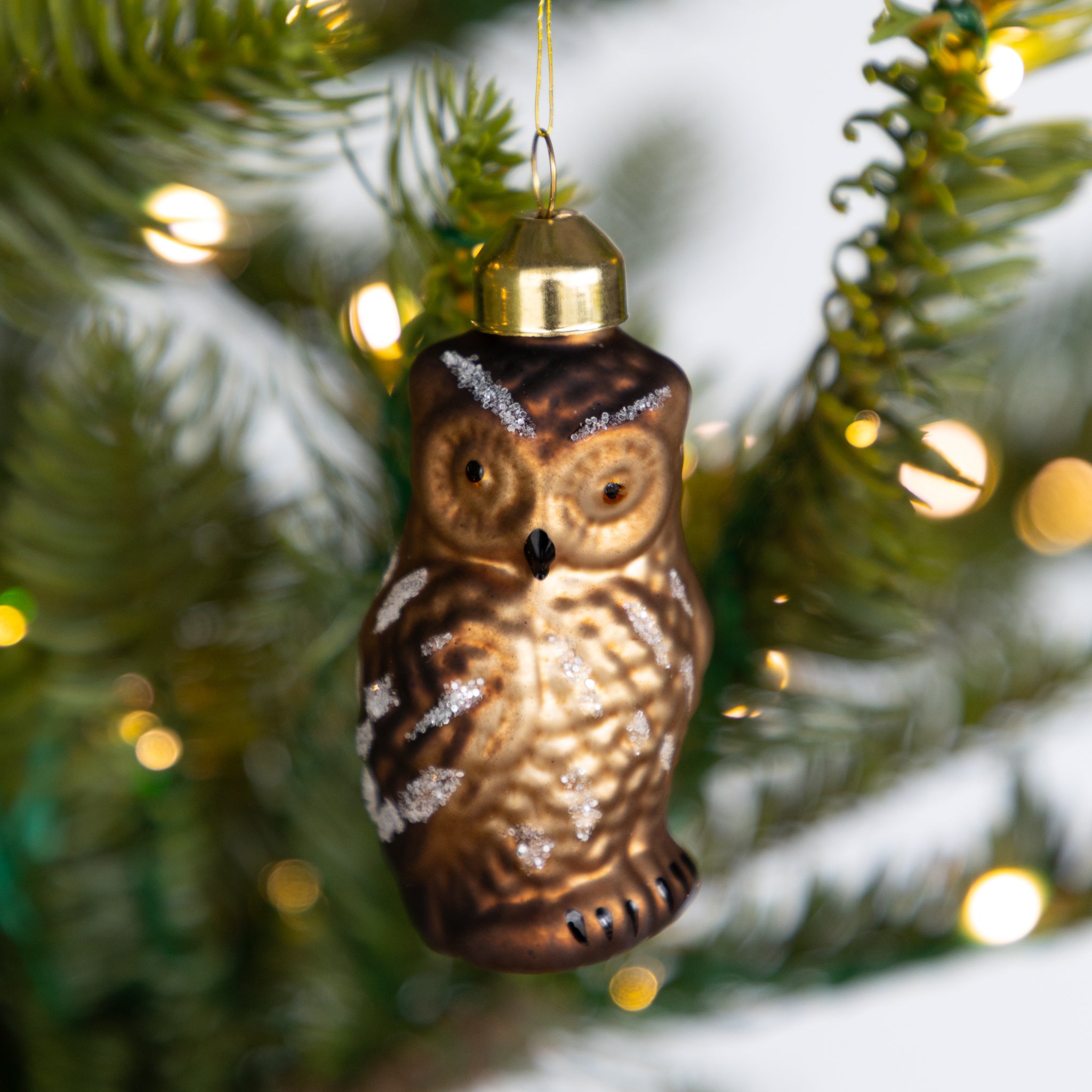 Decorative owl ornament hanging on a Christmas tree with lights in the background.