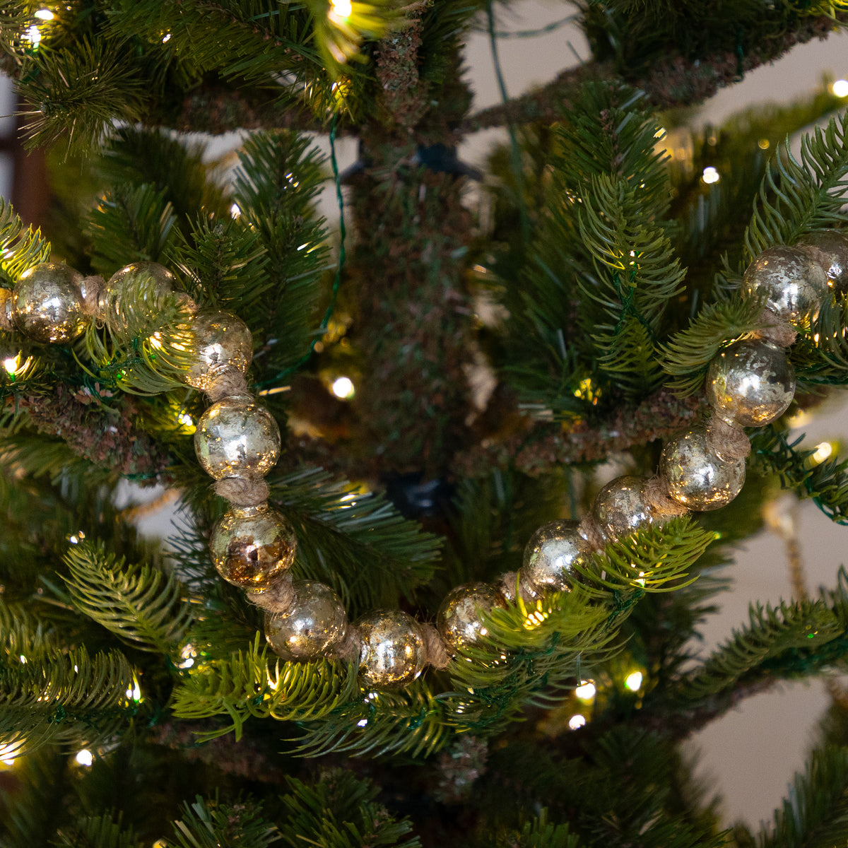 A close-up of a green artificial Christmas tree decorated with a strand of reflective silver bead garland and warm white string lights.