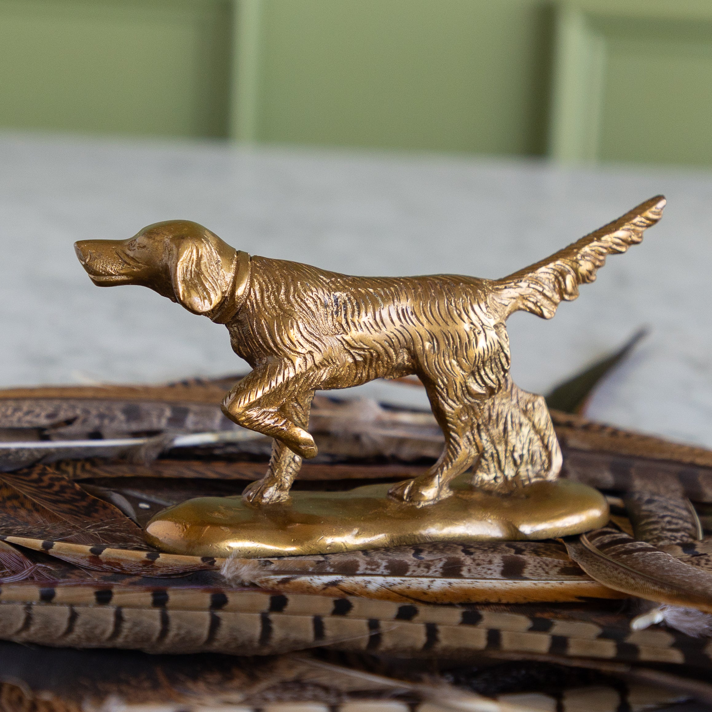 A small metallic dog figurine in a golden-bronze finish, posed standing on a base, photographed on a tray with feathers against a softly blurred indoor background.