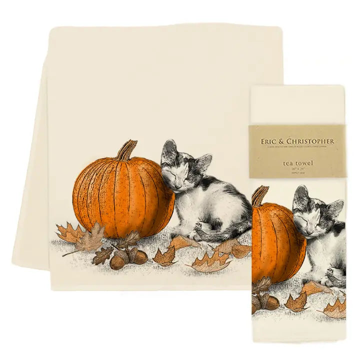 A cream-colored tea towel featuring an illustration of a black-and-white kitten next to an orange pumpkin with scattered autumn leaves, shown with its paper band packaging labeled Eric & CHRISTOPHER.