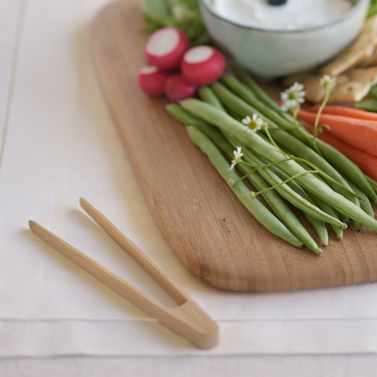 Wooden cutting board with green beans, carrots, radishes, and a bowl of dip on a light surface.