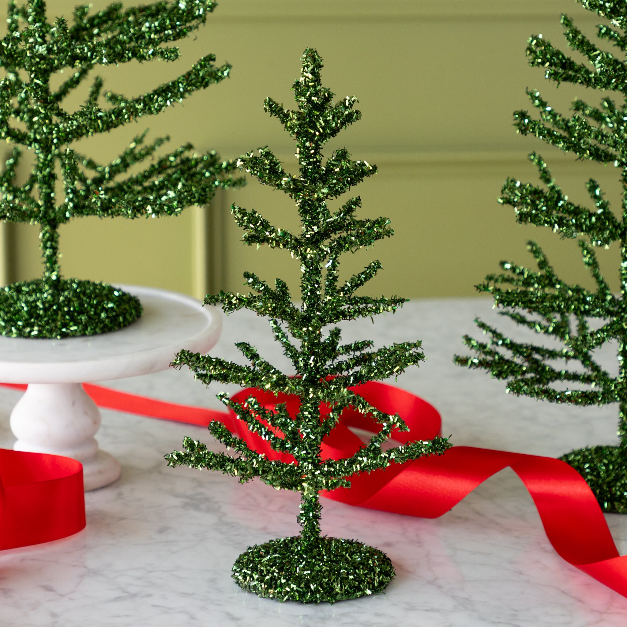 Three decorative green tinsel trees on a table with red ribbons against a green wall.