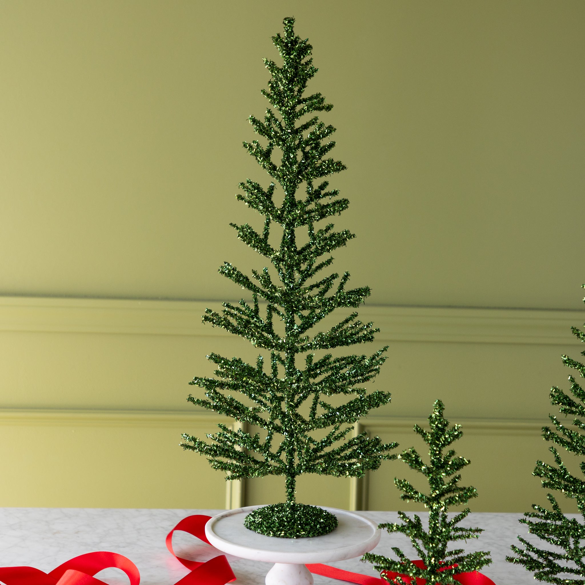 Large green artificial tinsel tree on a white surface with a red ribbon.