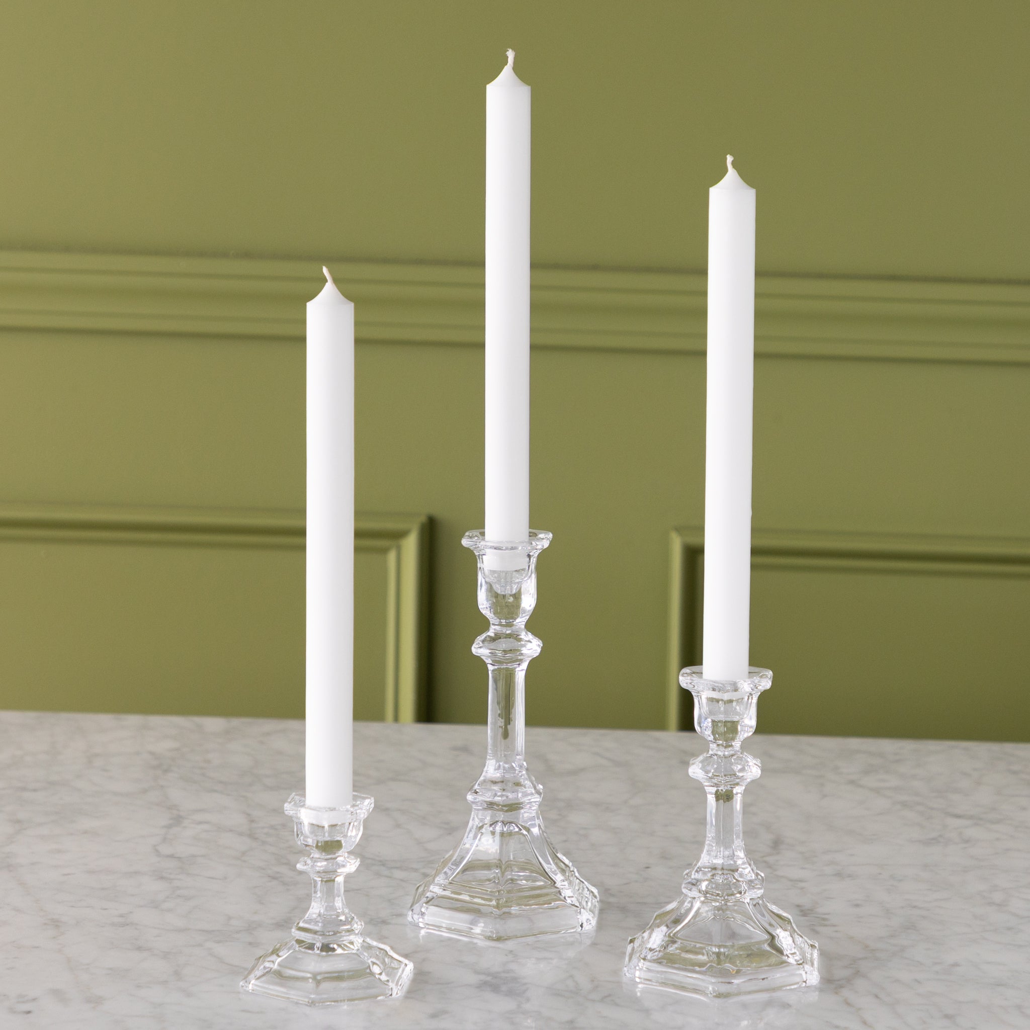 Three white candles in glass candlesticks on a marble surface with a green wall background.