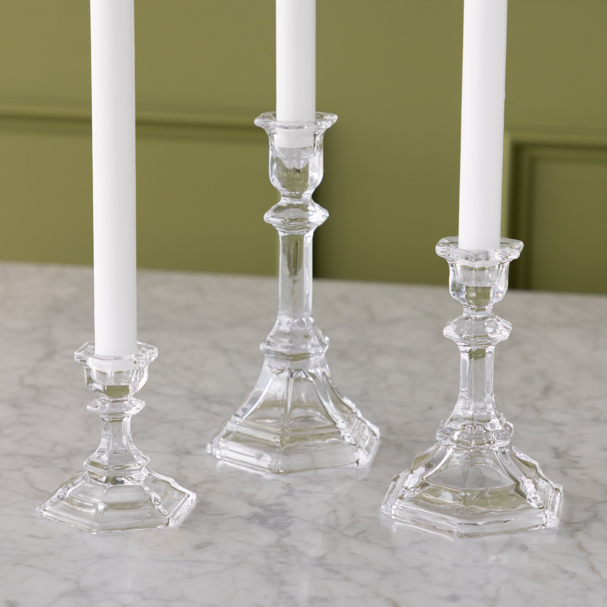 Three clear glass candlesticks with white candles on a marble surface against a green wall.
