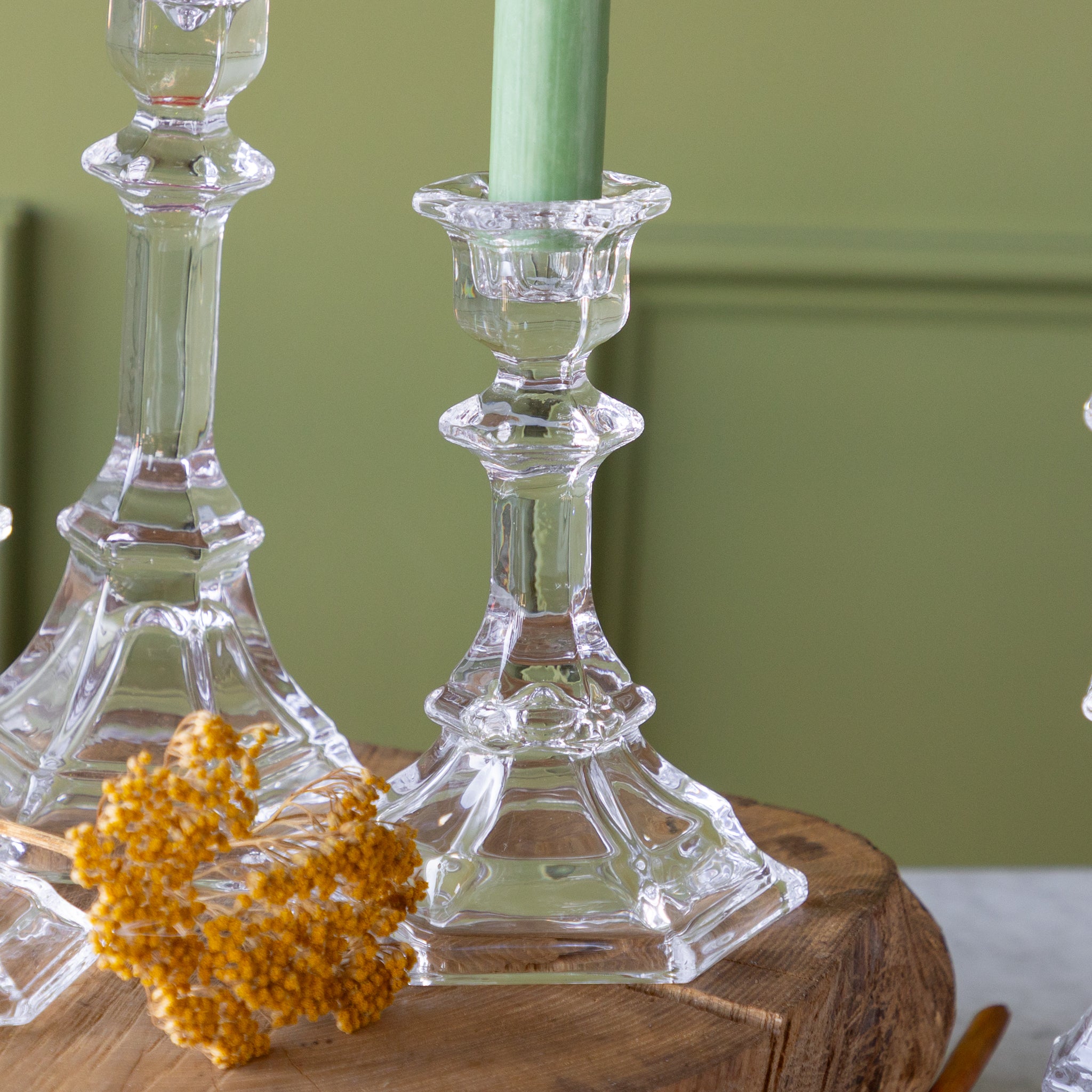 Clear glass candlesticks on a wooden surface with a green candle and decorative yellow leaves.