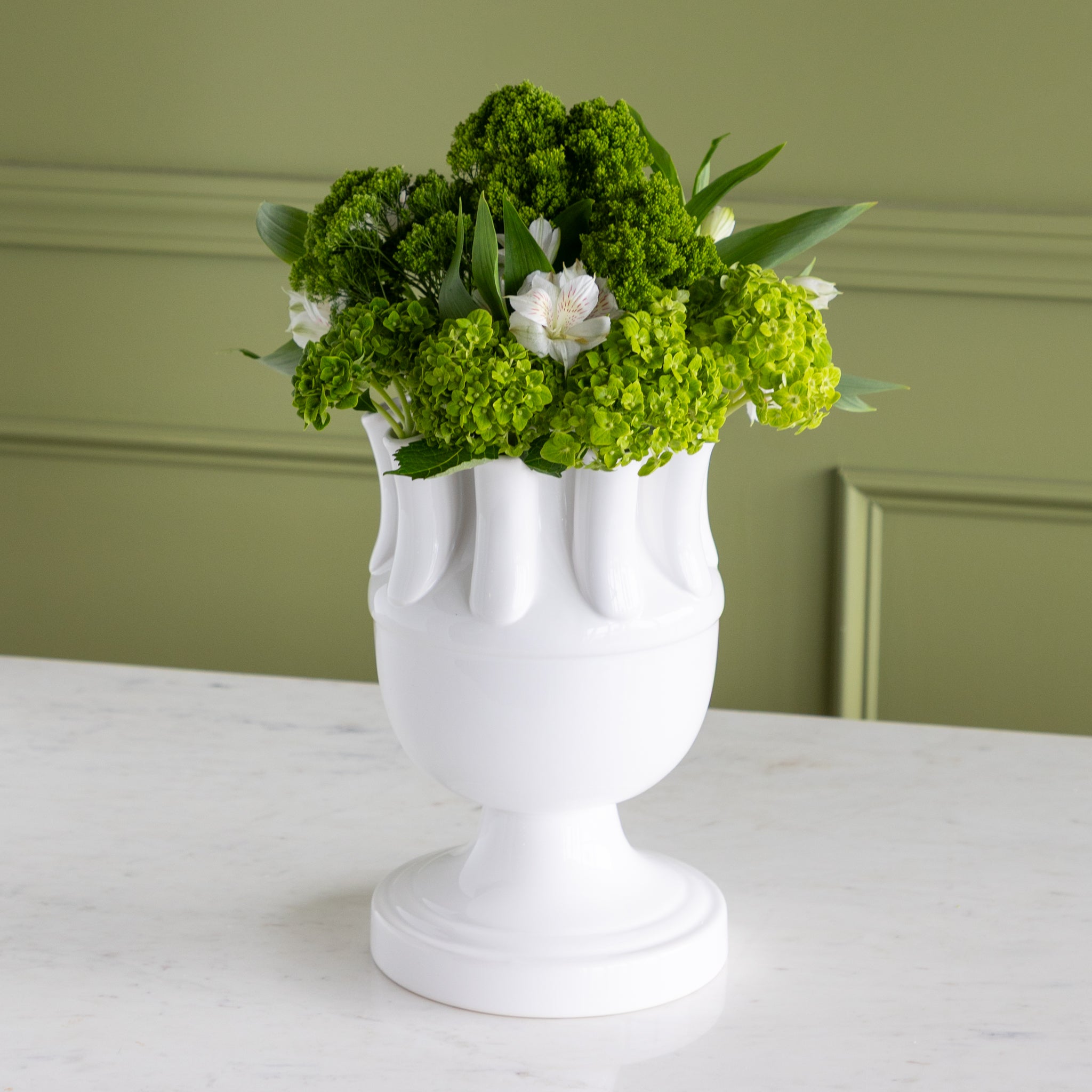White vase with green flowers against a green wall.