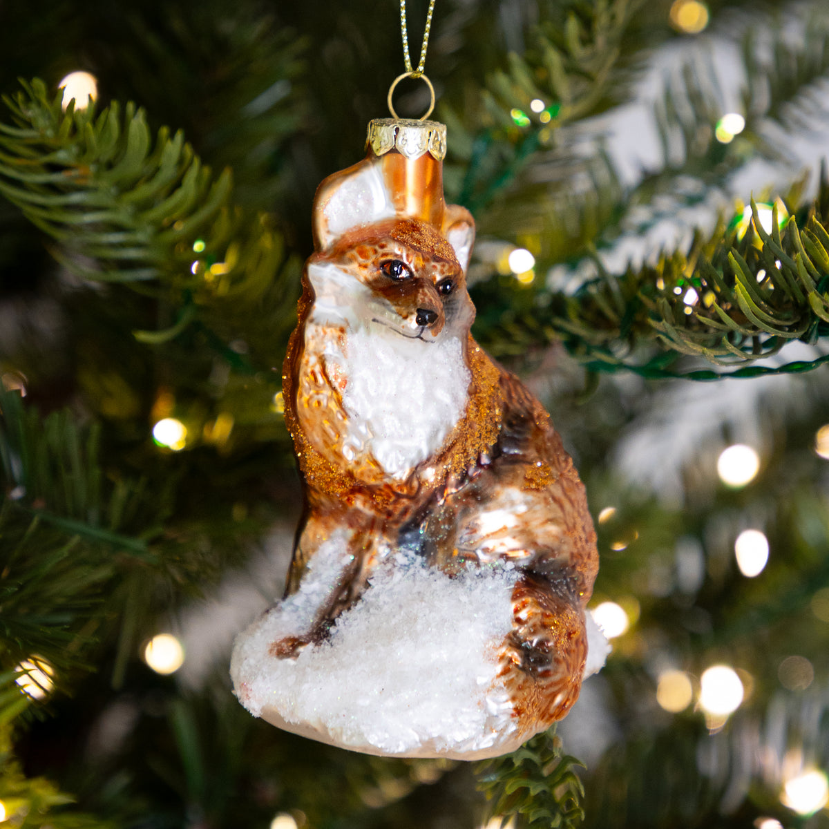 A hand-painted glass Christmas ornament shaped like a fox sitting on a snowy mound, hanging on a decorated Christmas tree with warm lights in the background.