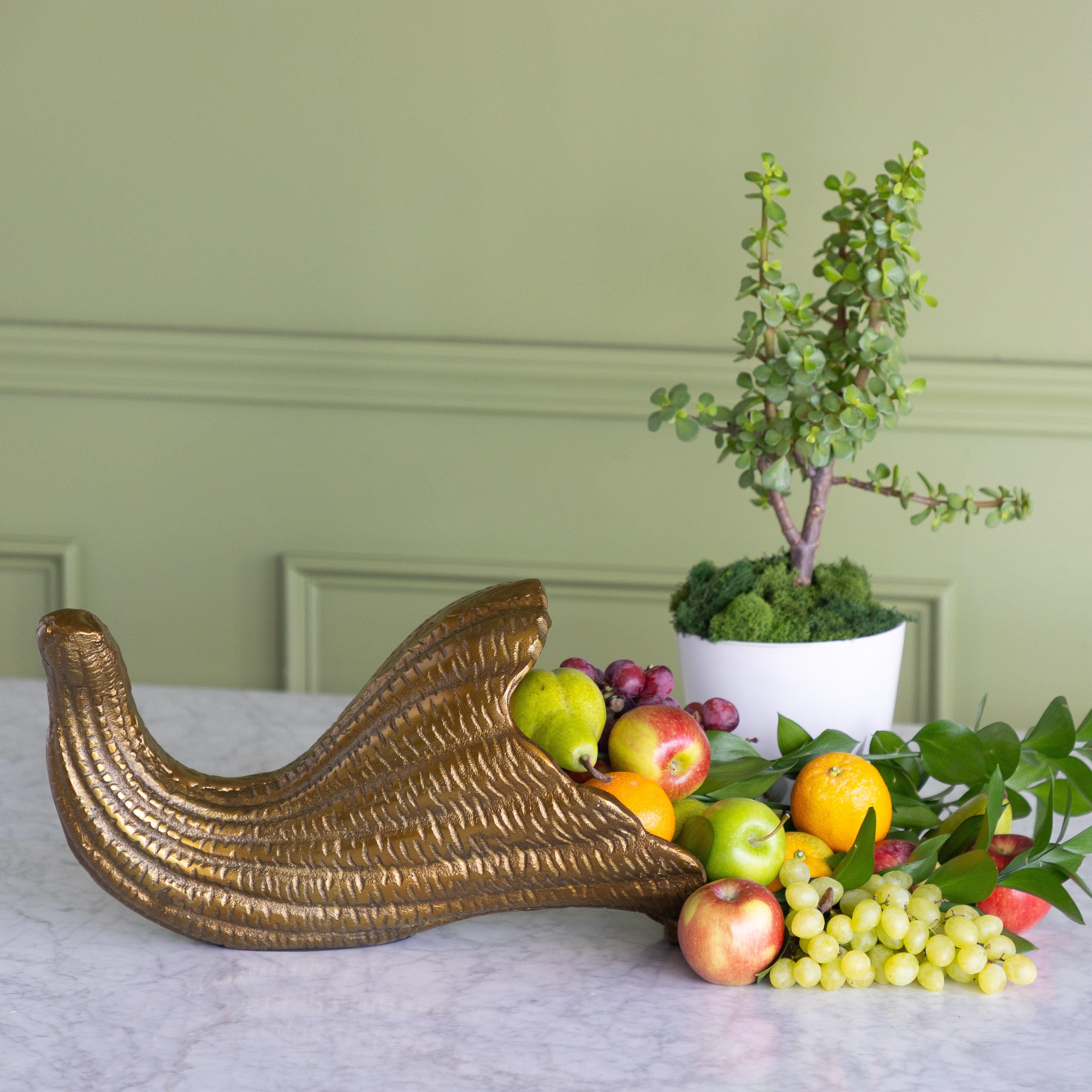 A decorative gold cornucopia-style bowl overflowing with fruit, placed on a marble surface in front of a potted plant against a sage green paneled wall.