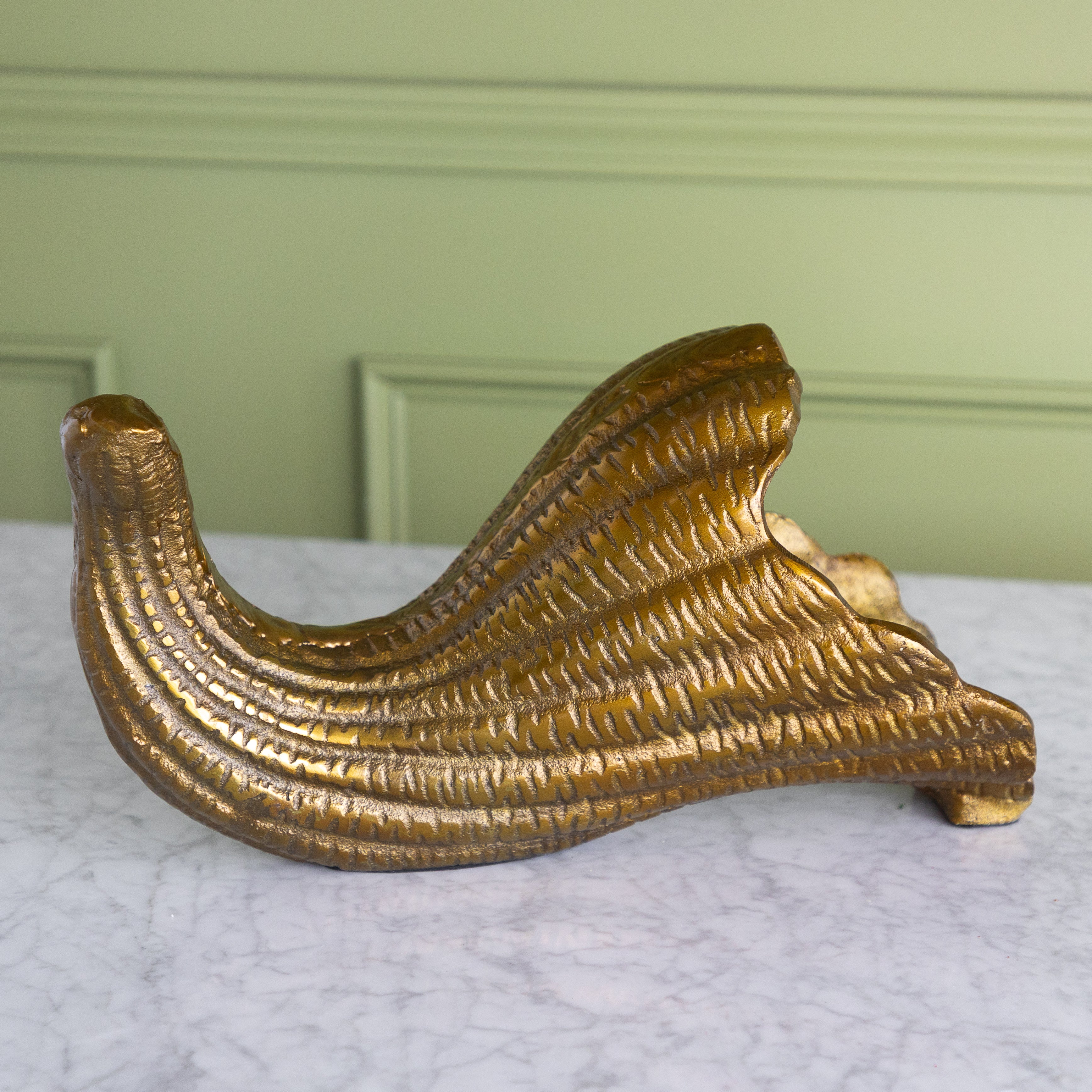A decorative gold-toned metal bracket or shelf support with a ribbed, wave-like textured surface, displayed on a white marble surface against a light green paneled wall.