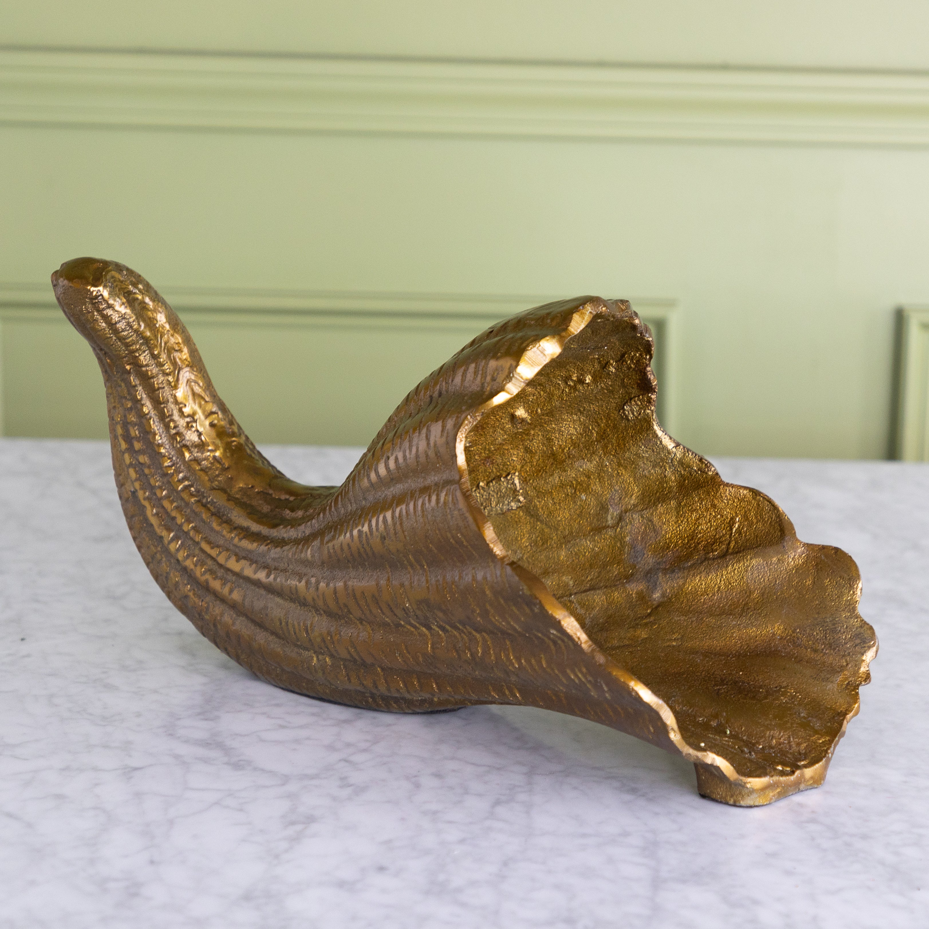 A decorative gold-toned horn-shaped bowl with a textured, ribbed surface and an irregular, scalloped rim, displayed on a white marble surface against a pale green paneled wall.