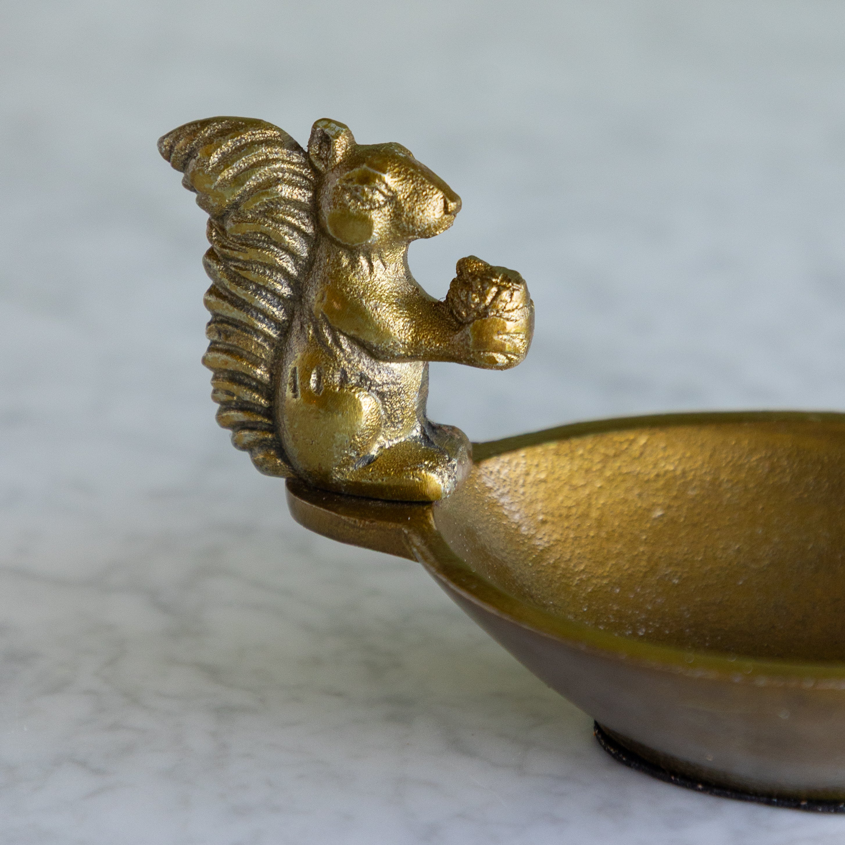 A close-up of a small metallic bowl with a sculpted squirrel handle holding an acorn, shown on a light marble-like surface.