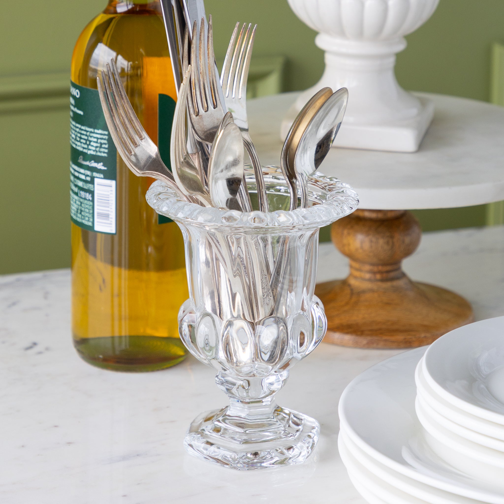 Silverware set in a clear glass holder on a table with a bottle and plates in the background.