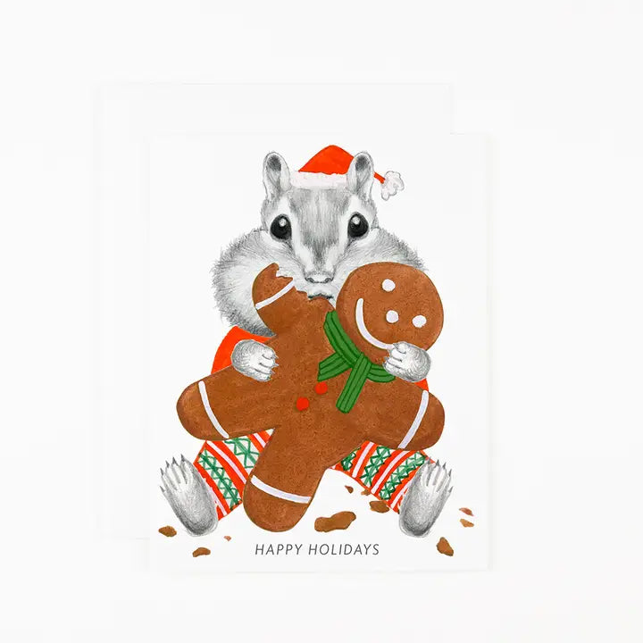 A festive holiday greeting card featuring a watercolor illustration of a gray squirrel wearing a red Santa hat and hugging a gingerbread man in red and green holiday clothing. The card is printed on white cardstock with &