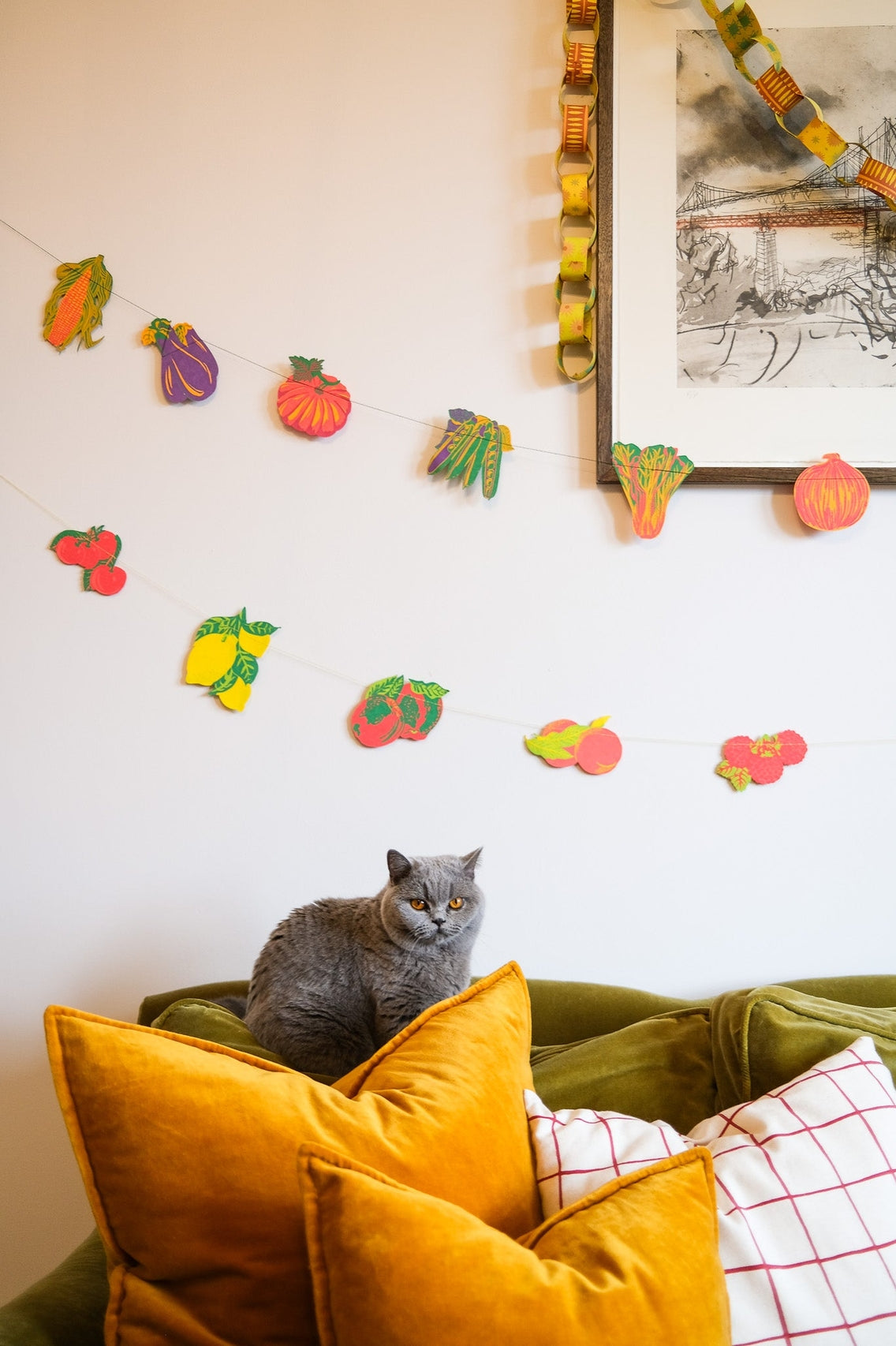Cat sitting on a green couch with colorful pillows, surrounded by decorative fruit and vegetable garland on the wall.
