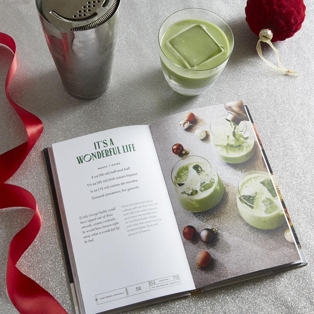 Open cookbook with green drinks and garnishes on a gray surface with a cocktail shaker and glass.