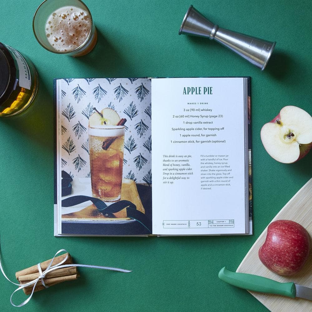 Open recipe book displaying an apple pie recipe with a glass of apple pie drink, apples, and cinnamon sticks on a green surface.