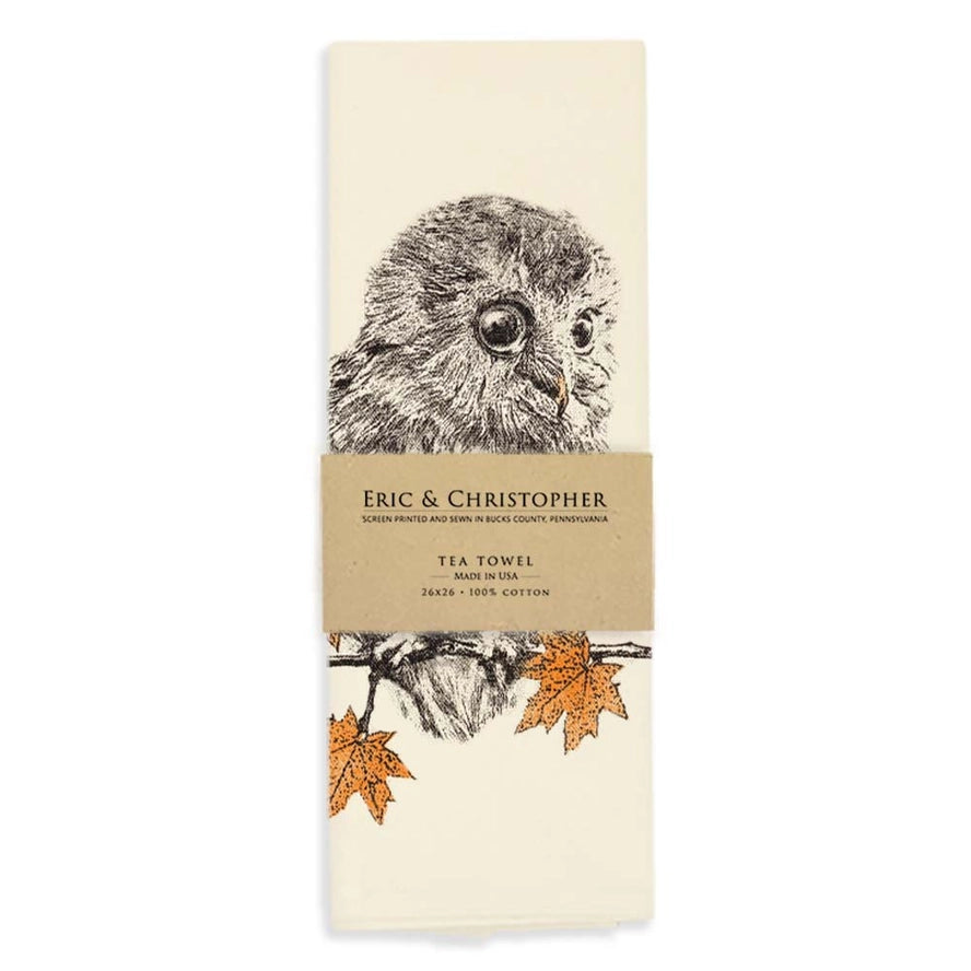 An off-white cotton tea towel featuring a detailed black ink illustration of an owl, with two orange autumn leaves at the bottom and a brown paper band wrapped around the middle. The band is printed with the brand &