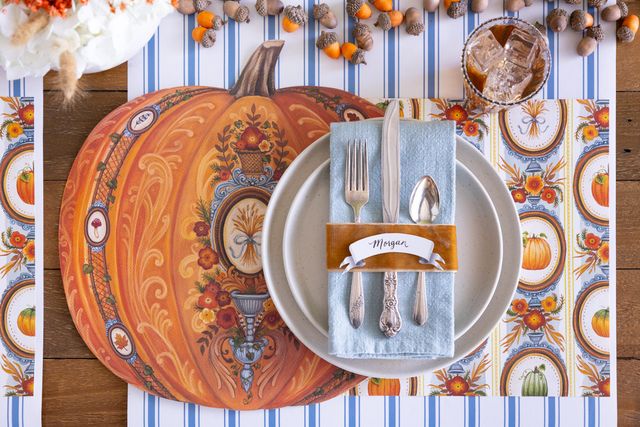 Thanksgiving table setting with pumpkin-themed placemat, plates, and cutlery on a striped tablecloth.