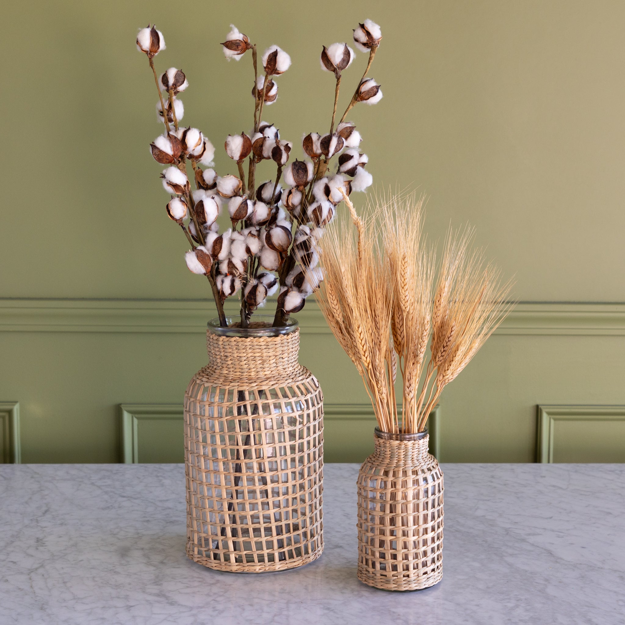 Two Rattan Wrapped Glass Vases with wheat stalks and cotton stems on a marble table against a green wall.