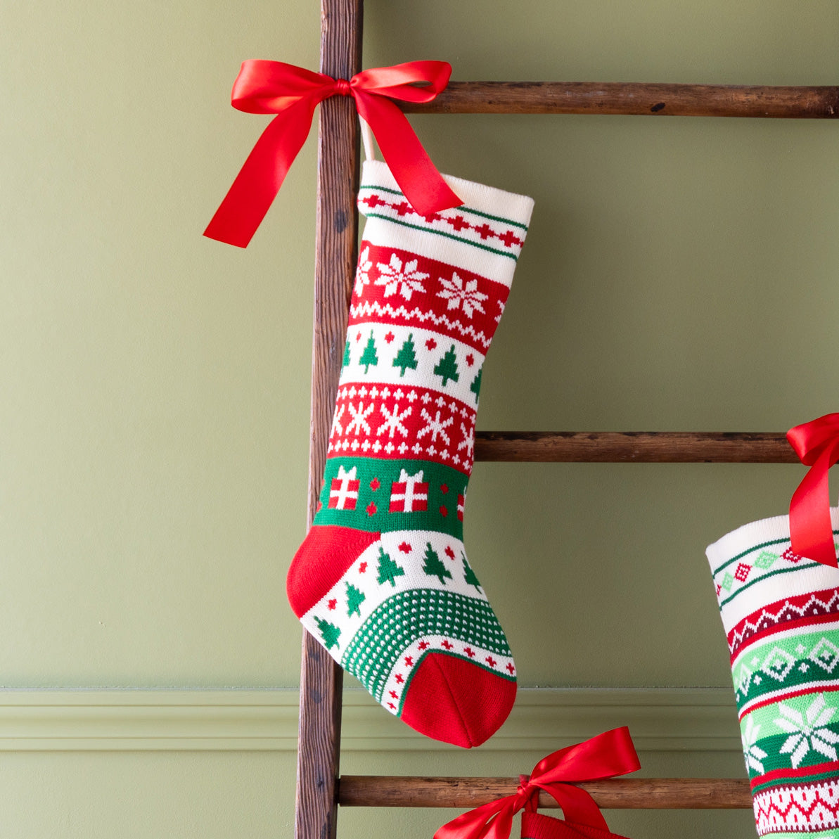 Decorative Christmas stocking with snowflakes, presents and trees on a wooden ladder against a green wall.