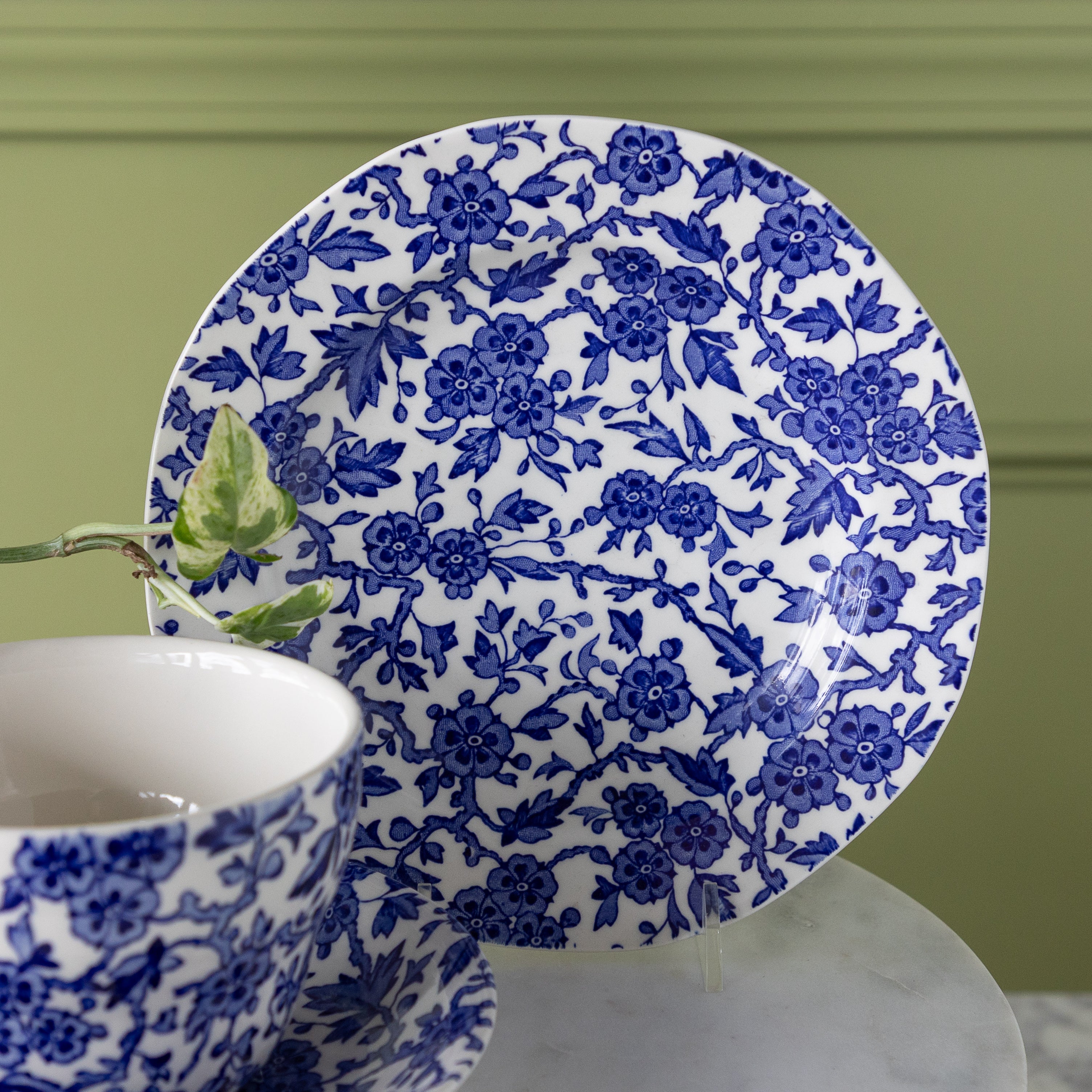 Burleigh Blue Arden Dinner Plate and Tea Cup with a blue floral pattern, against a green wall.
