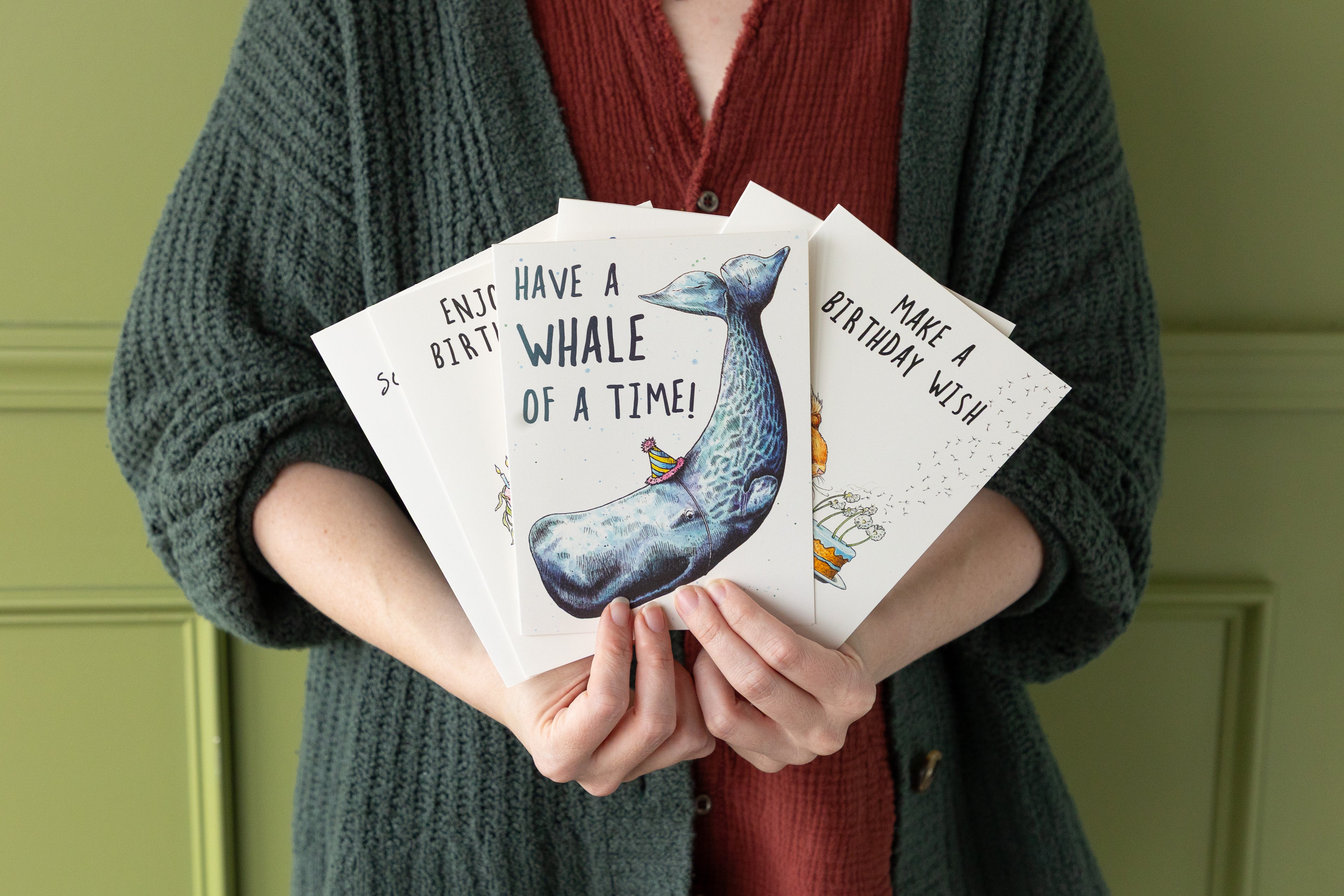Person holding birthday cards with whale illustrations against a green background.