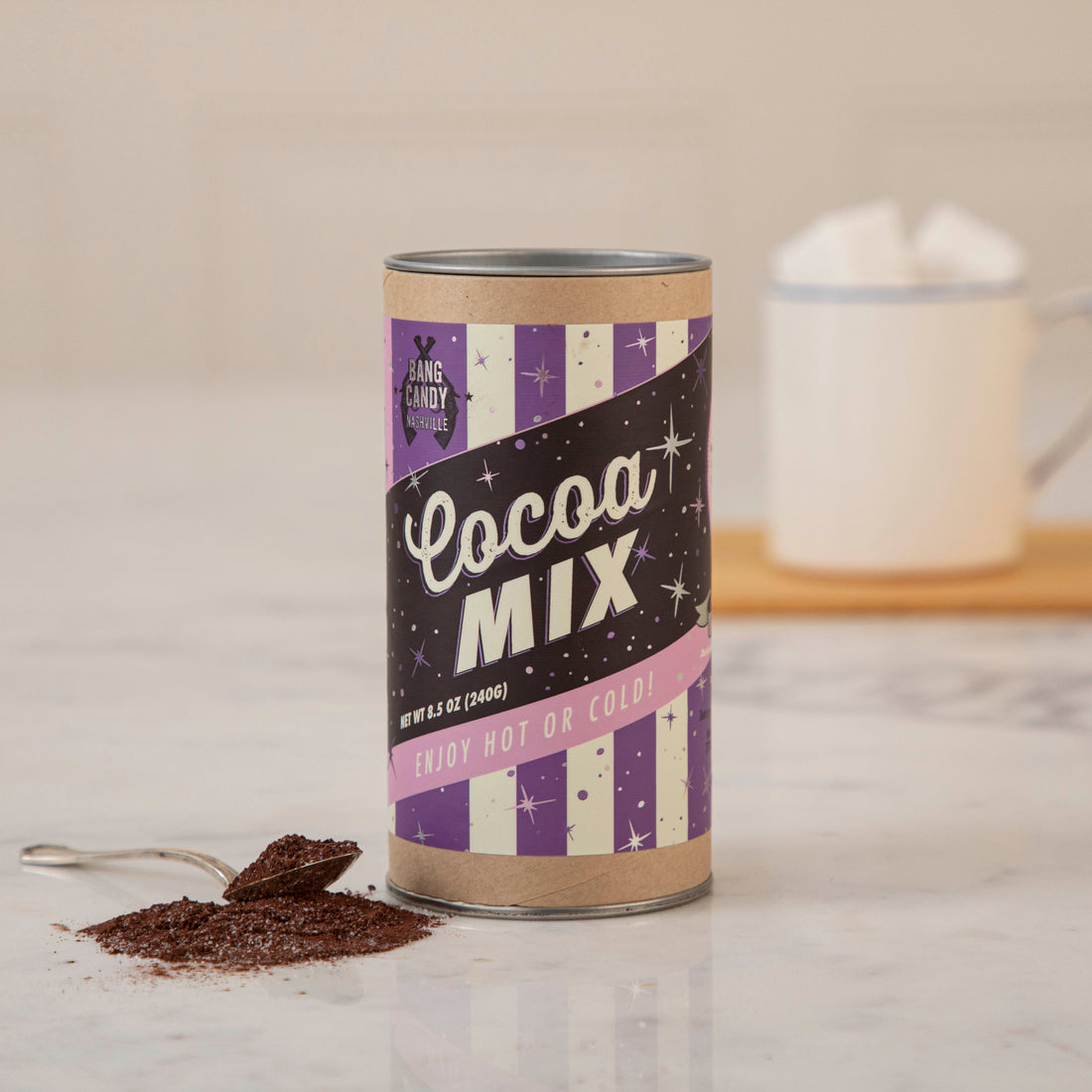A tin of Bang Candy Cocoa Mix next to a cup of coffee.