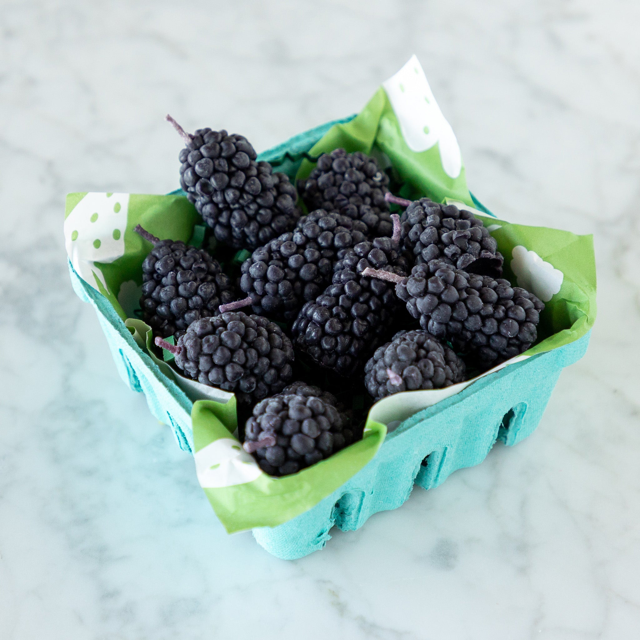 A blue carton filled with blackberry-shaped candles made from pure beeswax, on green paper grass, set on a white marble surface.