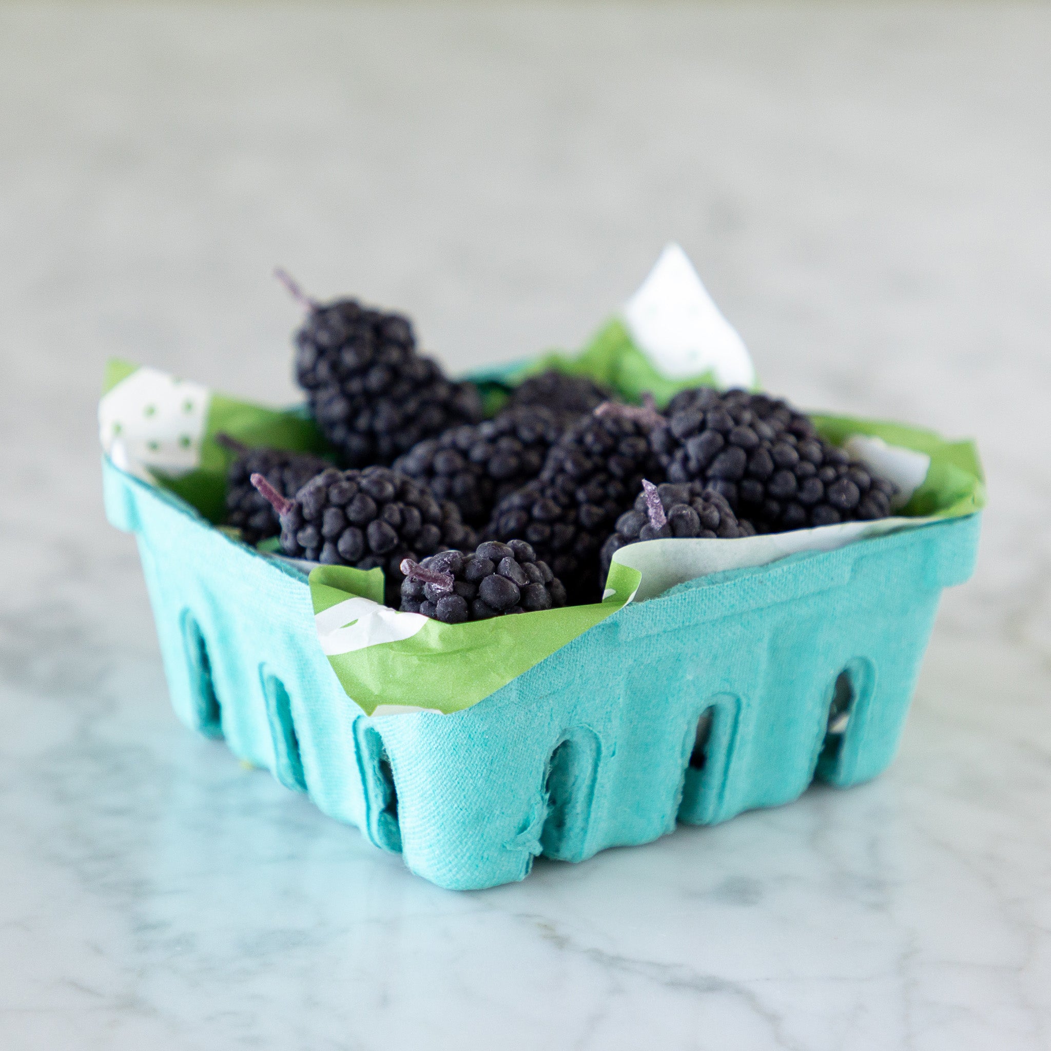 A blue carton filled with blackberry-shaped candles made from pure beeswax, on green paper grass, set on a white marble surface.