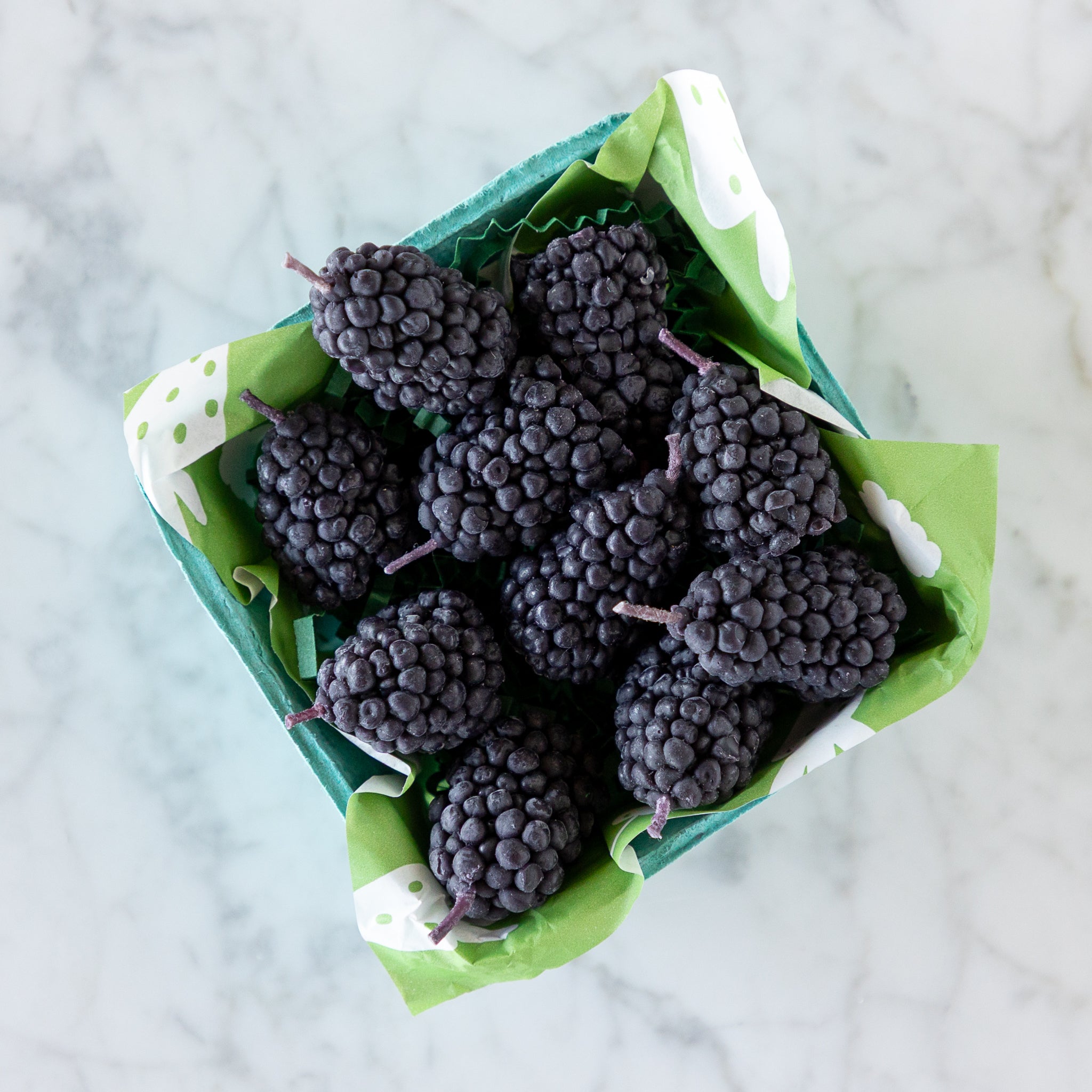 A blue carton filled with blackberry-shaped candles made from pure beeswax, on green paper grass, set on a white marble surface.