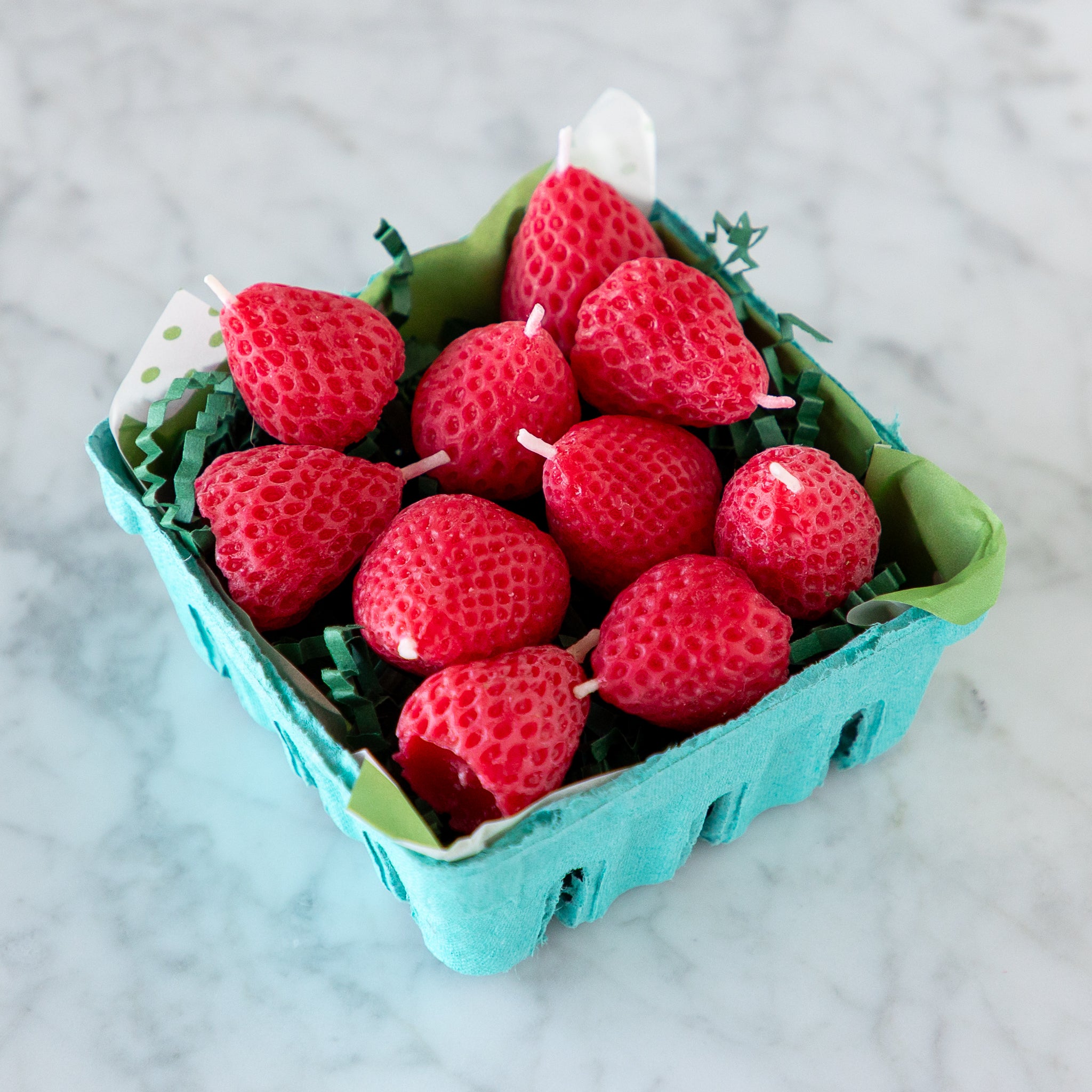 A blue carton filled with ten red, strawberry-shaped candles made from pure beeswax, on green paper grass, set on a white marble surface.