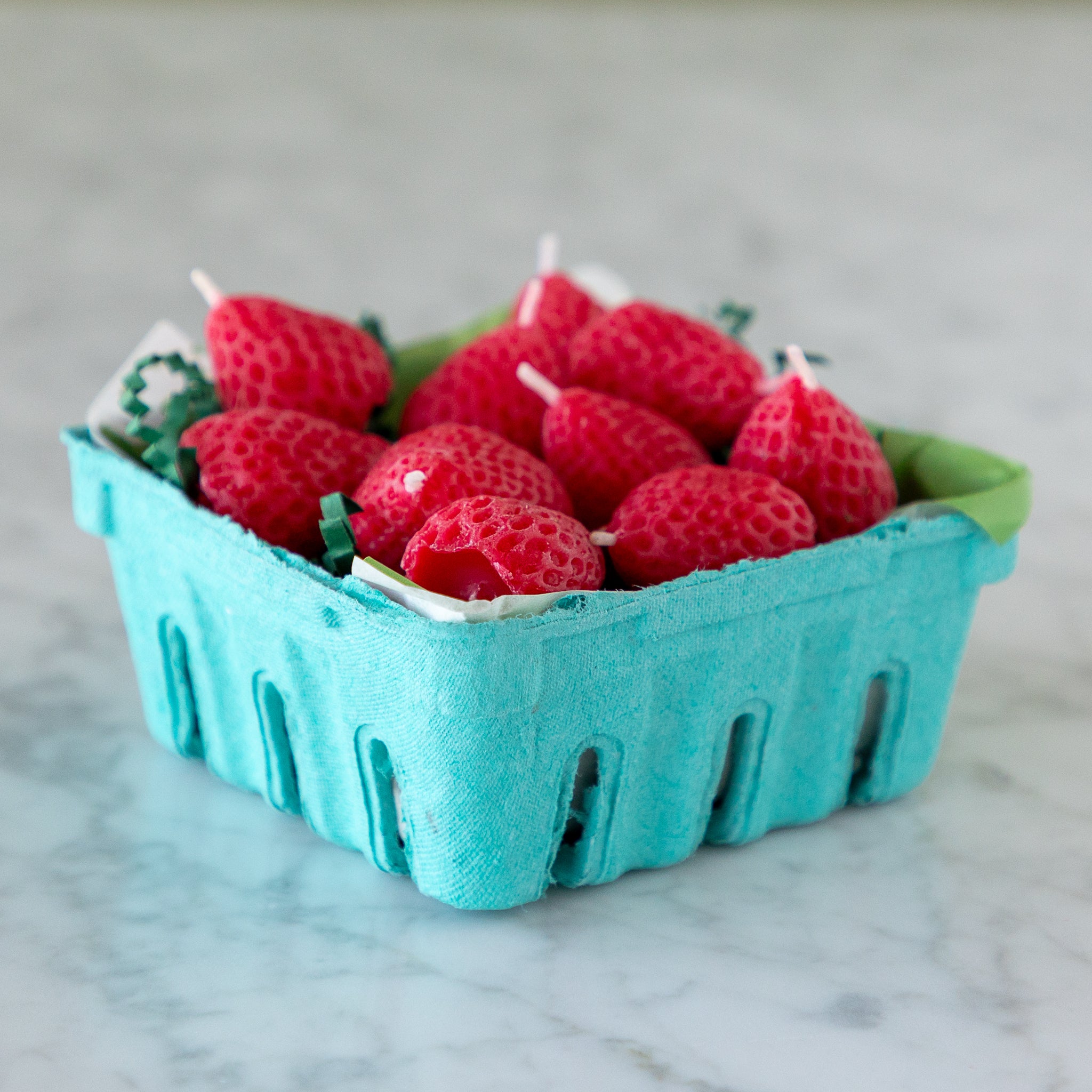A blue carton filled with ten red, strawberry-shaped candles made from pure beeswax, on green paper grass, set on a white marble surface.