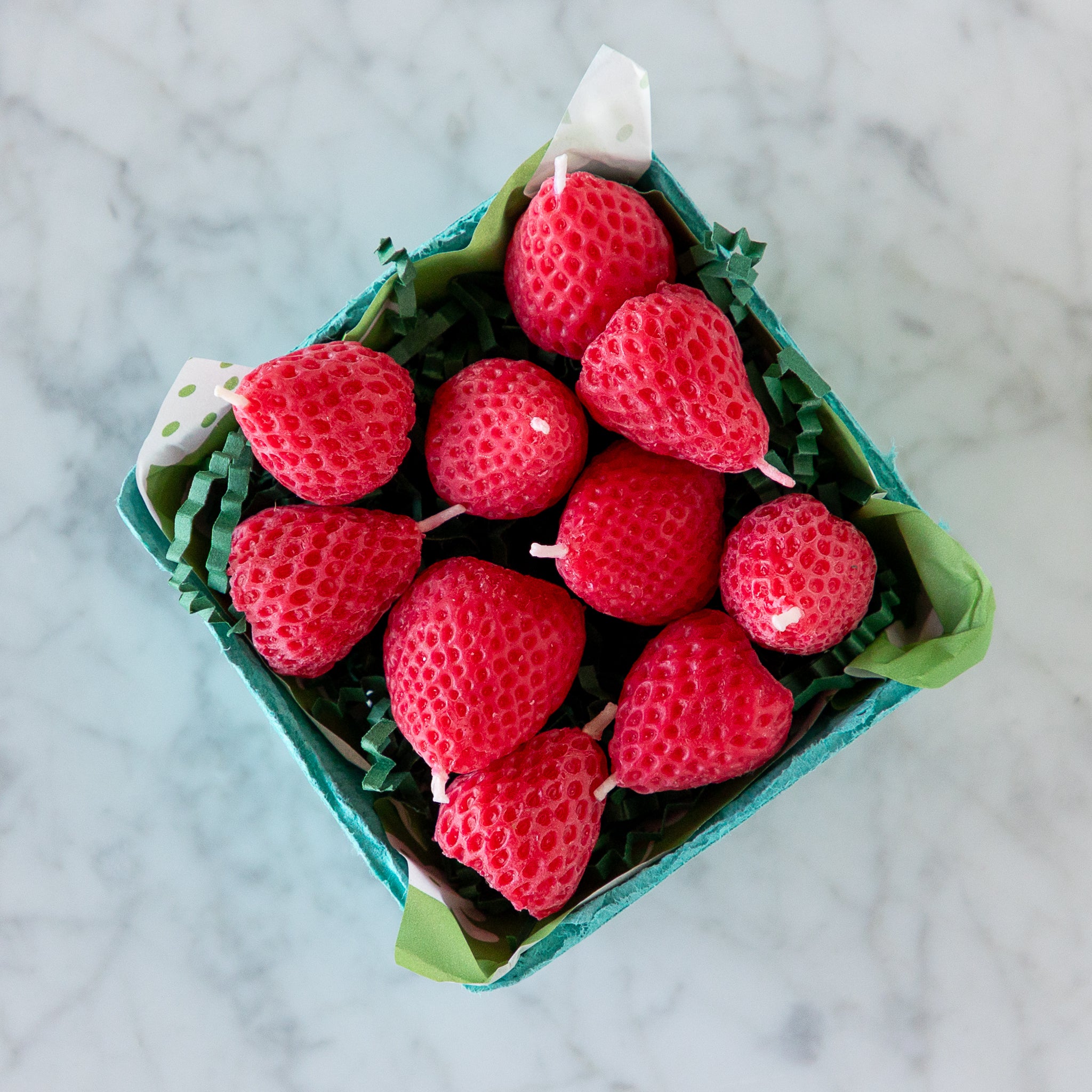A blue carton filled with ten red, strawberry-shaped candles made from pure beeswax, on green paper grass, set on a white marble surface.
