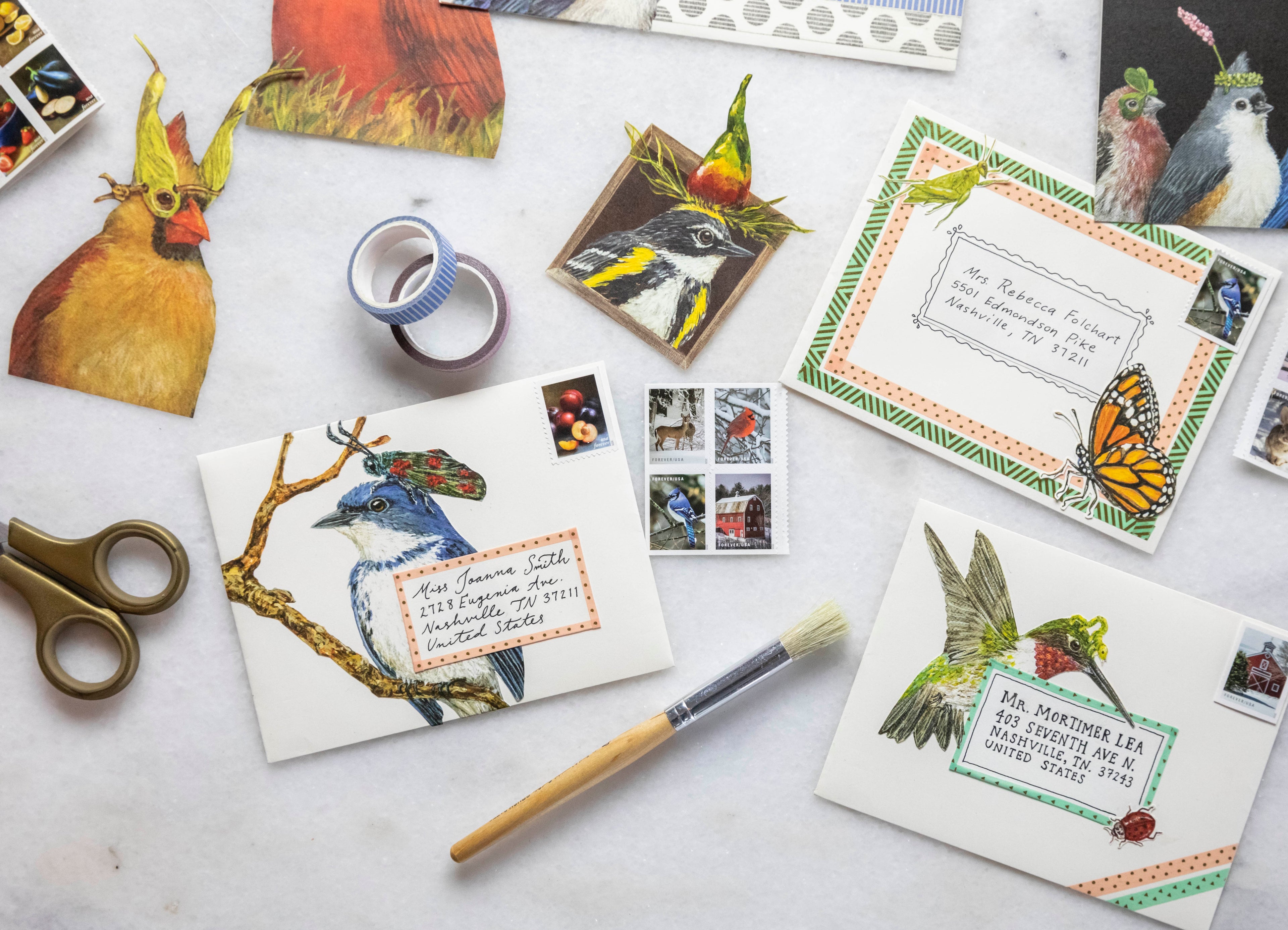 A top-down view of a white table covered in craft materials, featuring envelopes decorated with washi tape and artwork cut from various Vicki Sawyer greeting cards.