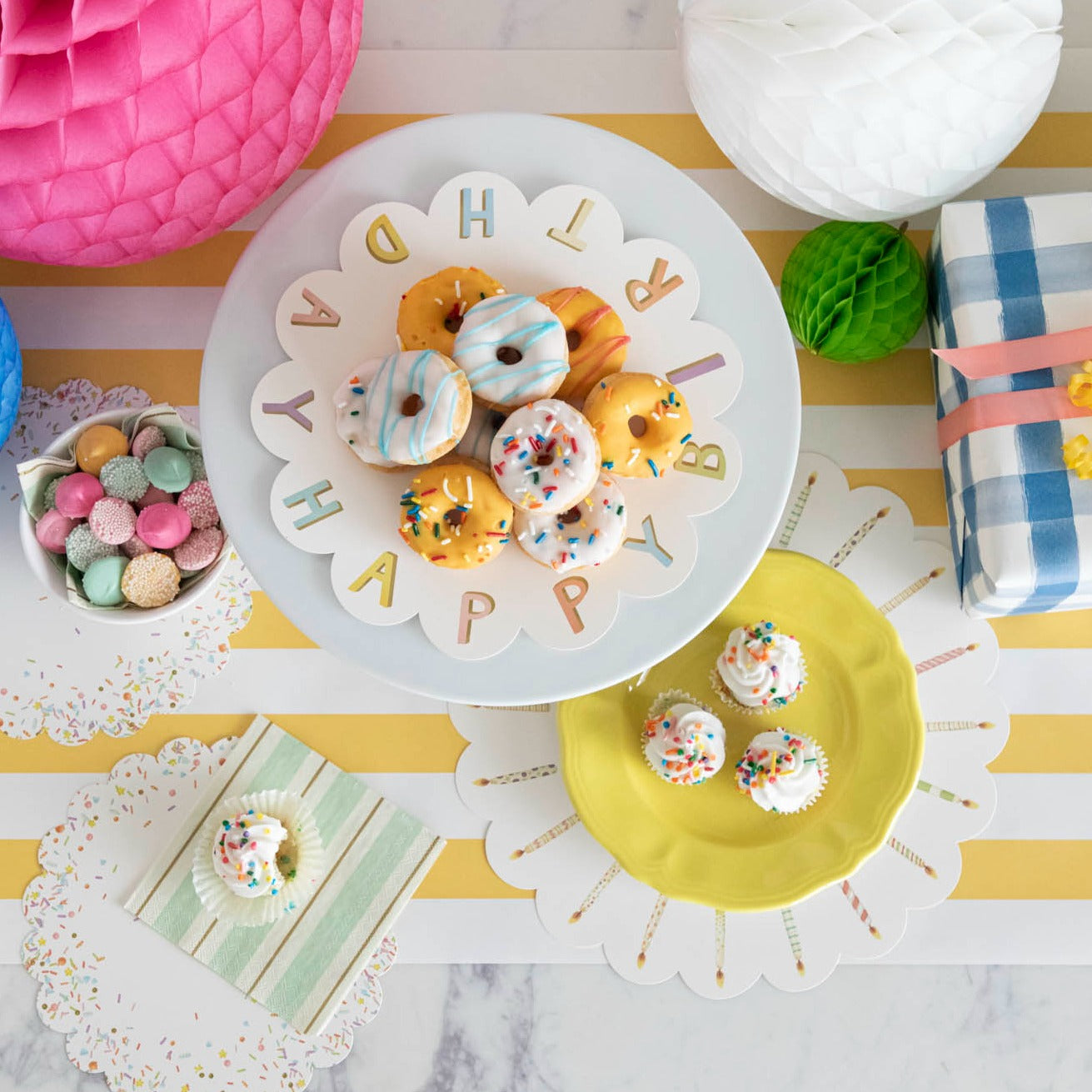 A plate of Happy Birthday Serving Papers donuts and cupcakes on a cake stand on a table. Brand: Hester & Cook.