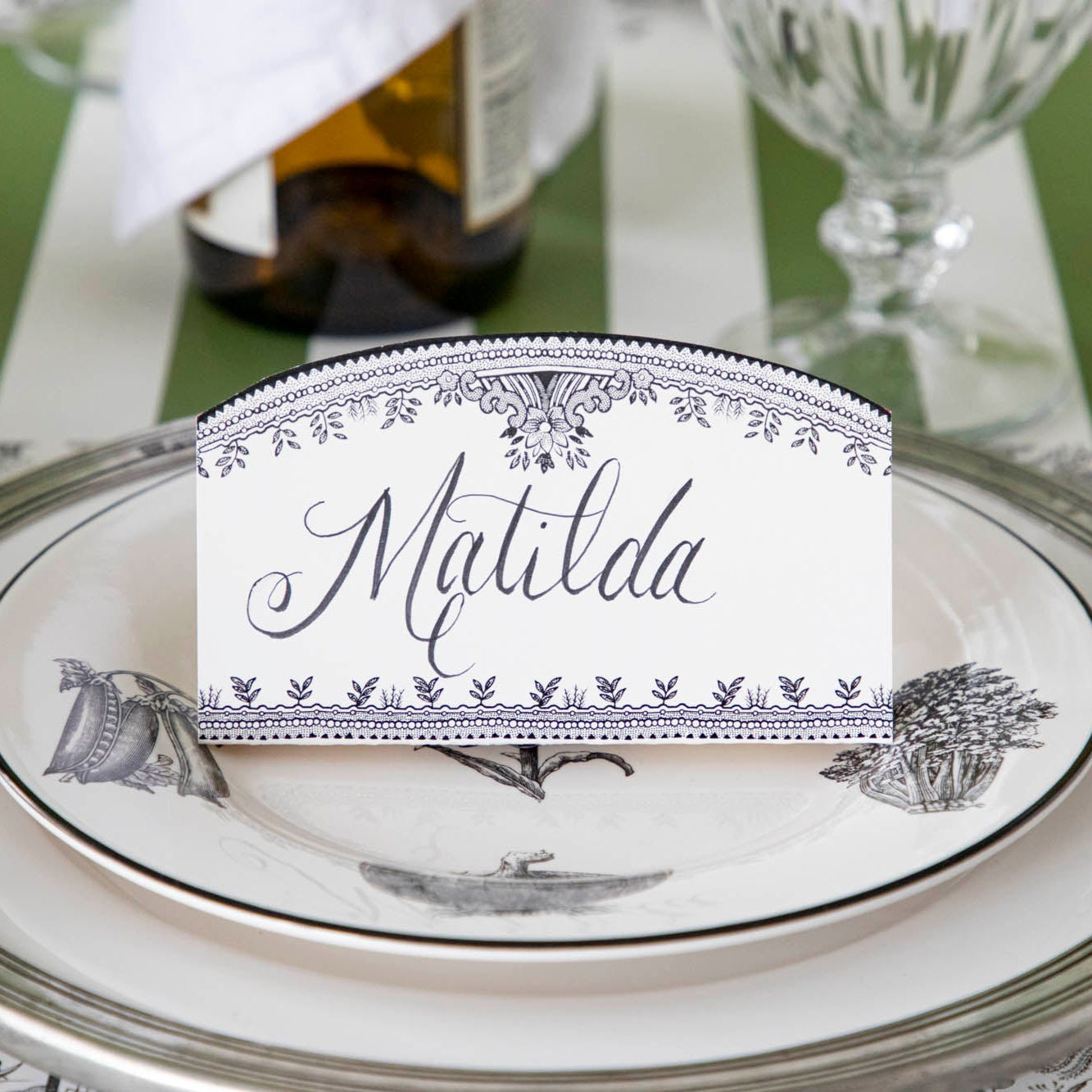 Black Willow Place Card with "Matilda" written on it, resting on dinner plates on a place setting.