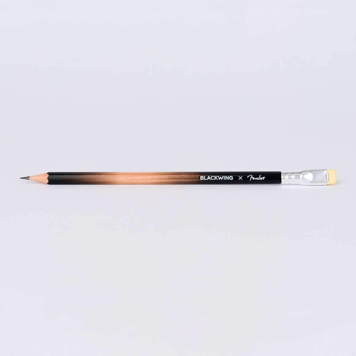 A Fender x BLACKWING collaboration pencil with a black-to-copper gradient barrel, a black grip, and a yellow eraser, shown horizontally on a light gray background.