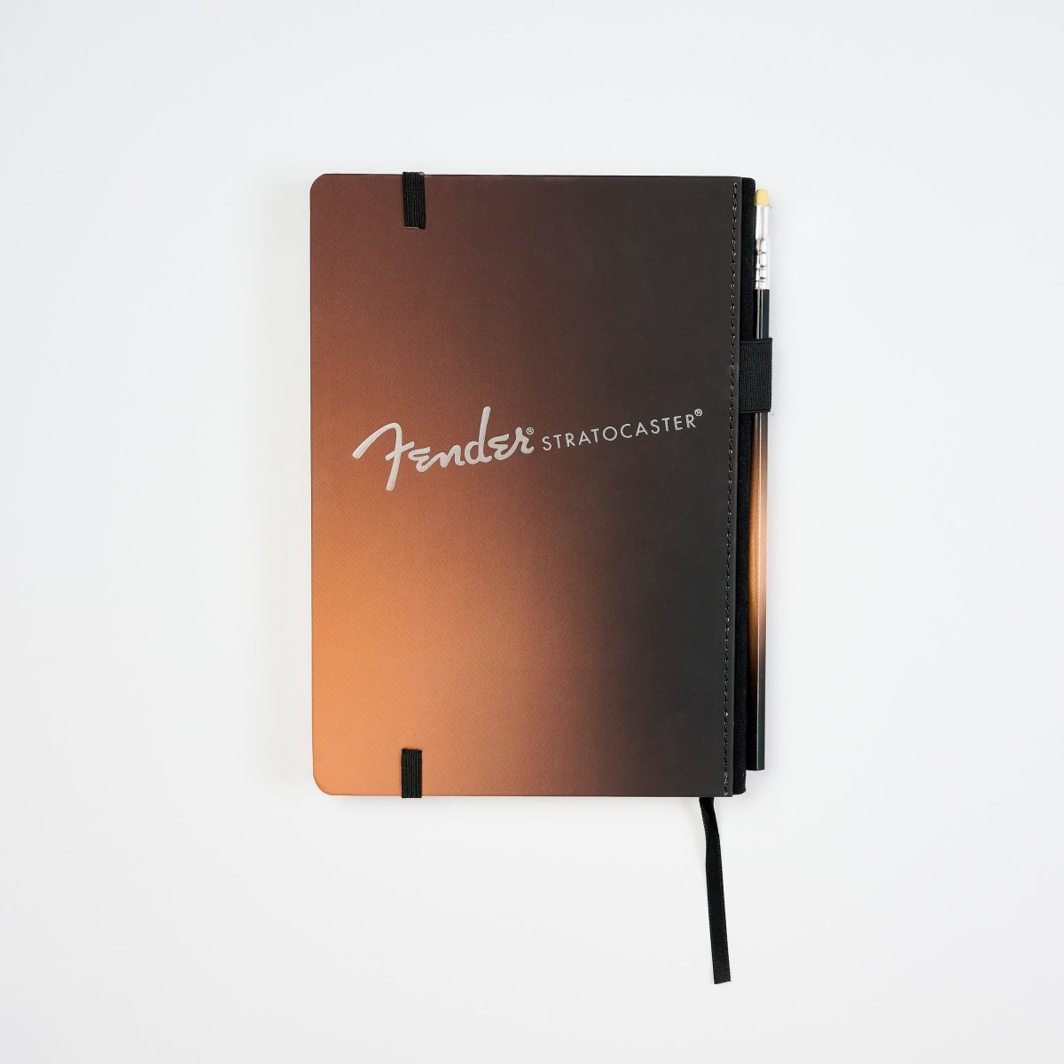 A Fender Stratocaster hardcover notebook with a black-to-orange gradient cover, featuring the white Fender logo and a black pen attached to the side, set against a plain white background.