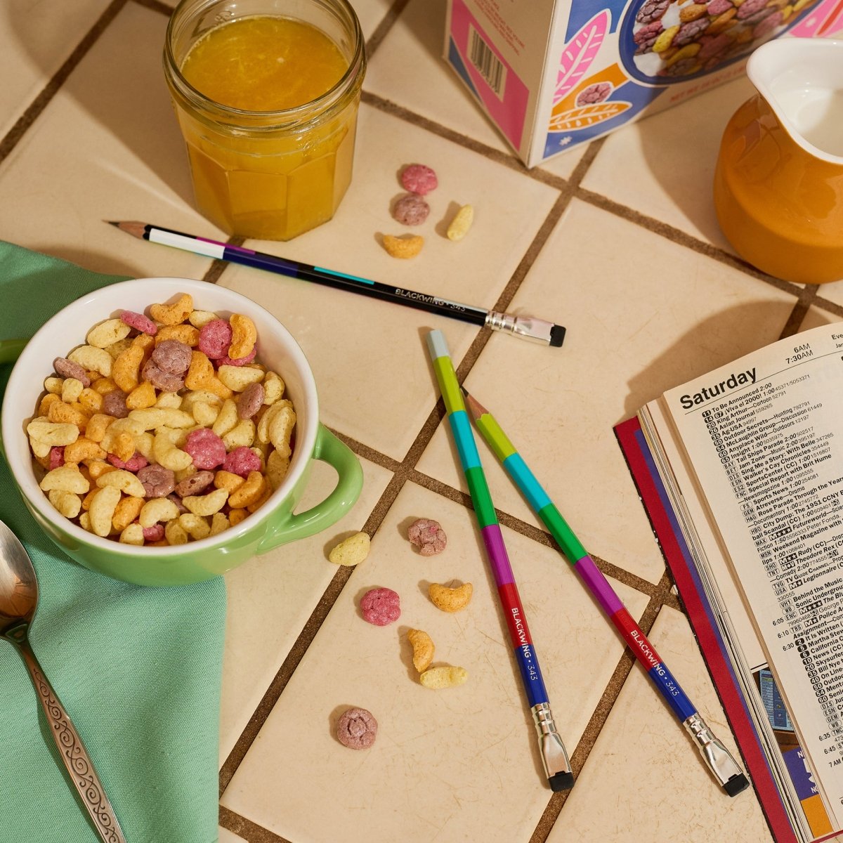 Blackwing pencils laying scattered on a table with breakfast food near them and a tv guide open.