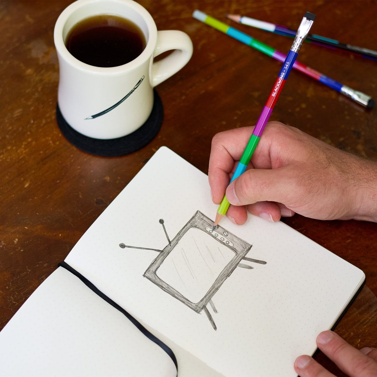 Person drawing in a notebook with colored pencils, next to a mug of coffee on a wooden table.