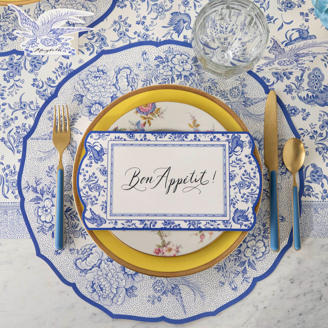 An elegant blue and white place setting featuring a Blue Regal Peacock Table Card with &quot;Bon Appetit&quot; written on it in beautiful cursive resting on the plate, from above.