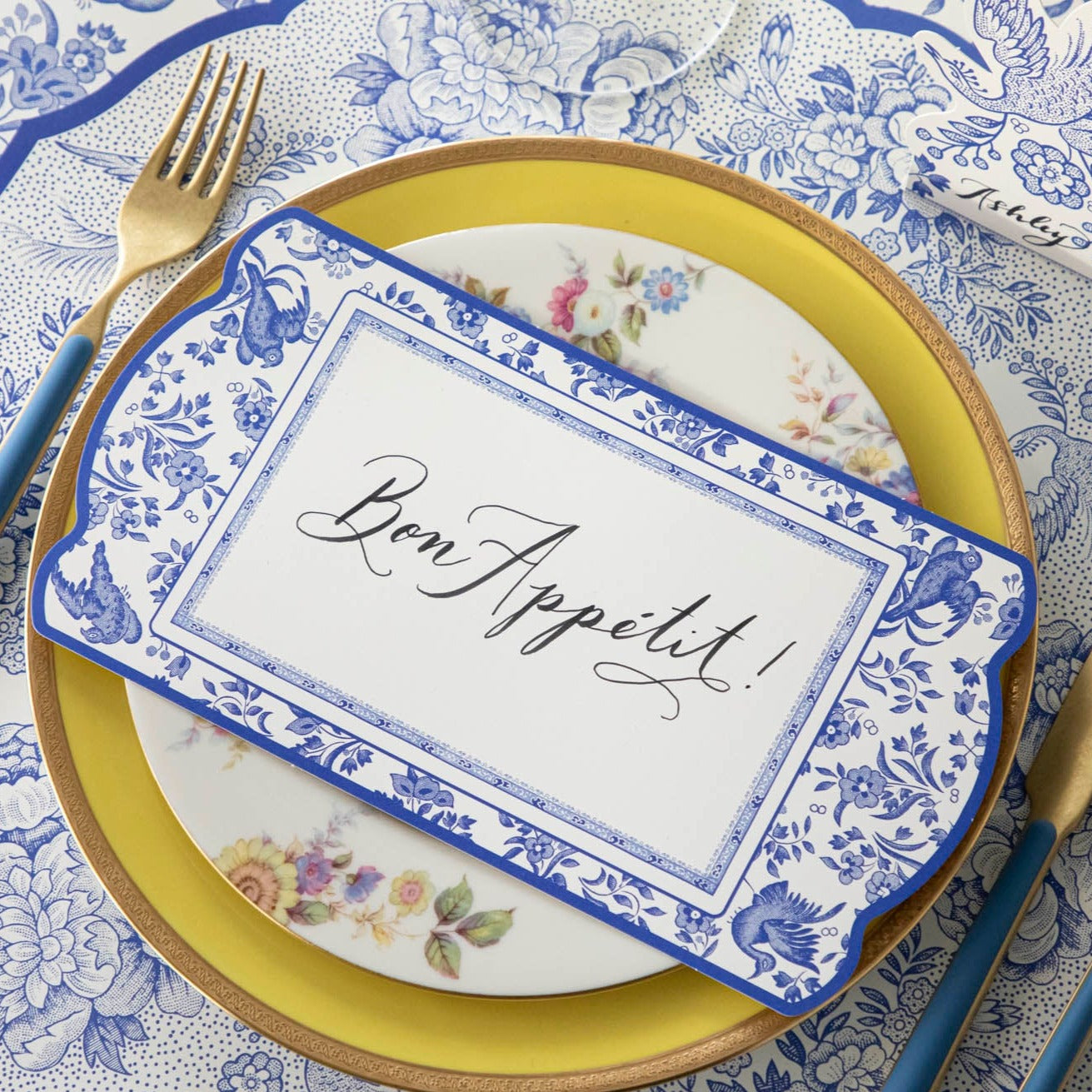 An elegant blue and white place setting featuring a Blue Regal Peacock Table Card with &quot;Bon Appetit&quot; written on it in beautiful cursive resting on the plate.