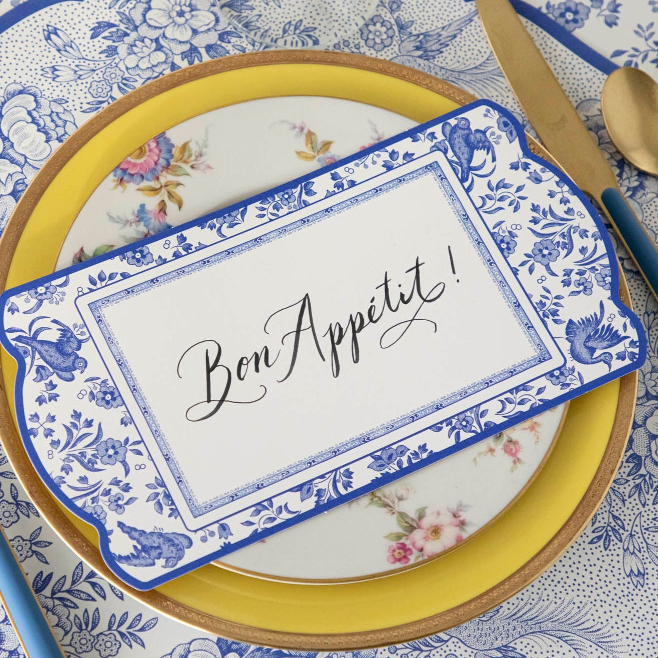 An elegant blue and white place setting featuring a Blue Regal Peacock Table Card with &quot;Bon Appetit&quot; written on it in beautiful cursive resting on the plate.