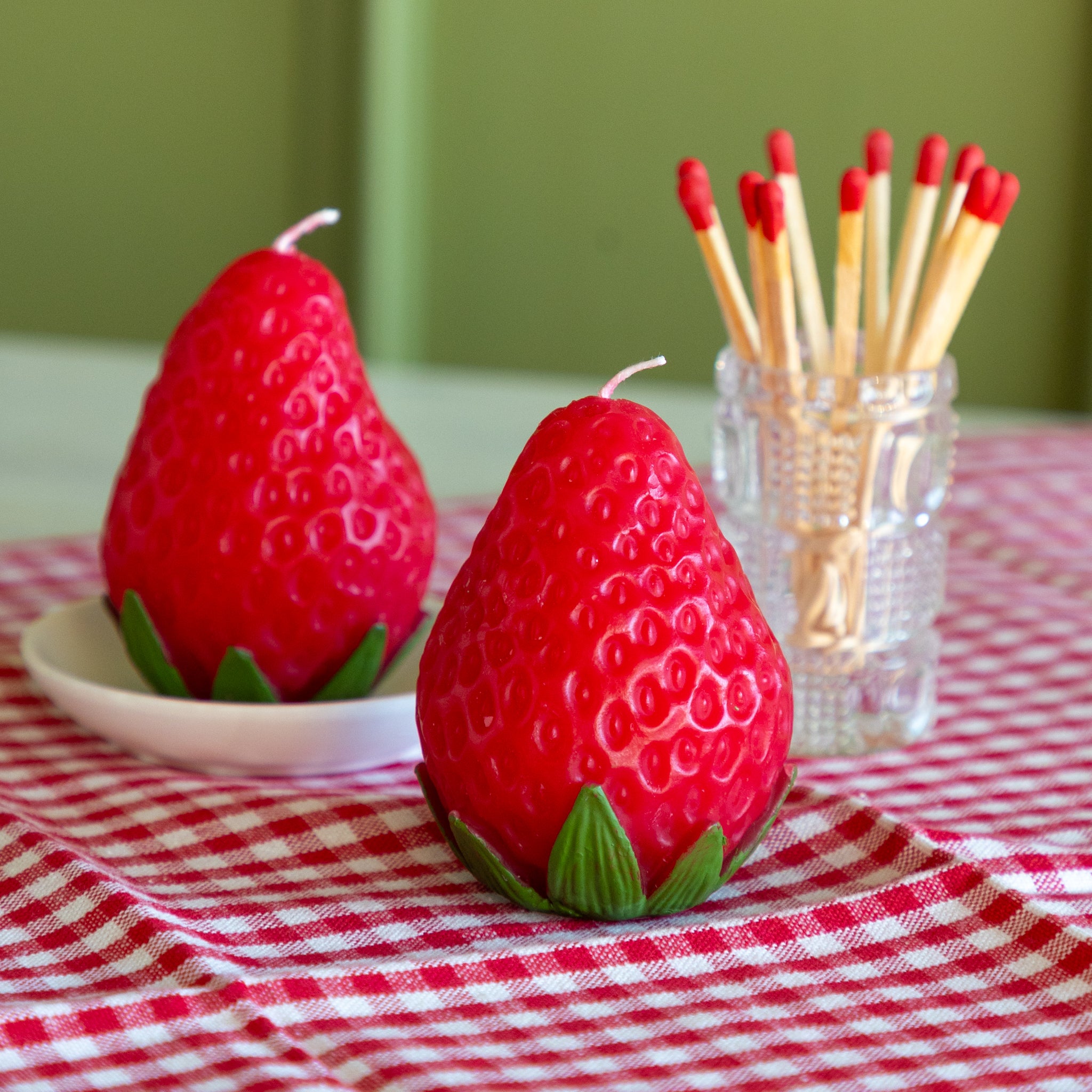 Two strawberry-shaped candles and a cup of matches on a table with a red and white checkered tablecloth.