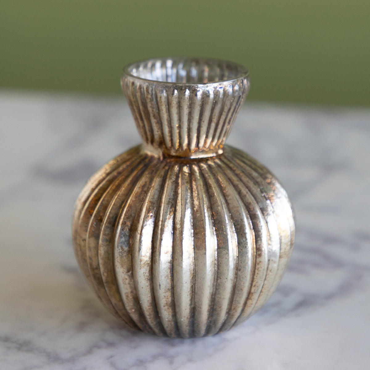 A small decorative vase with a ribbed, fluted texture in a metallic silver finish, resting on a white marble surface with a soft-focus olive green background.